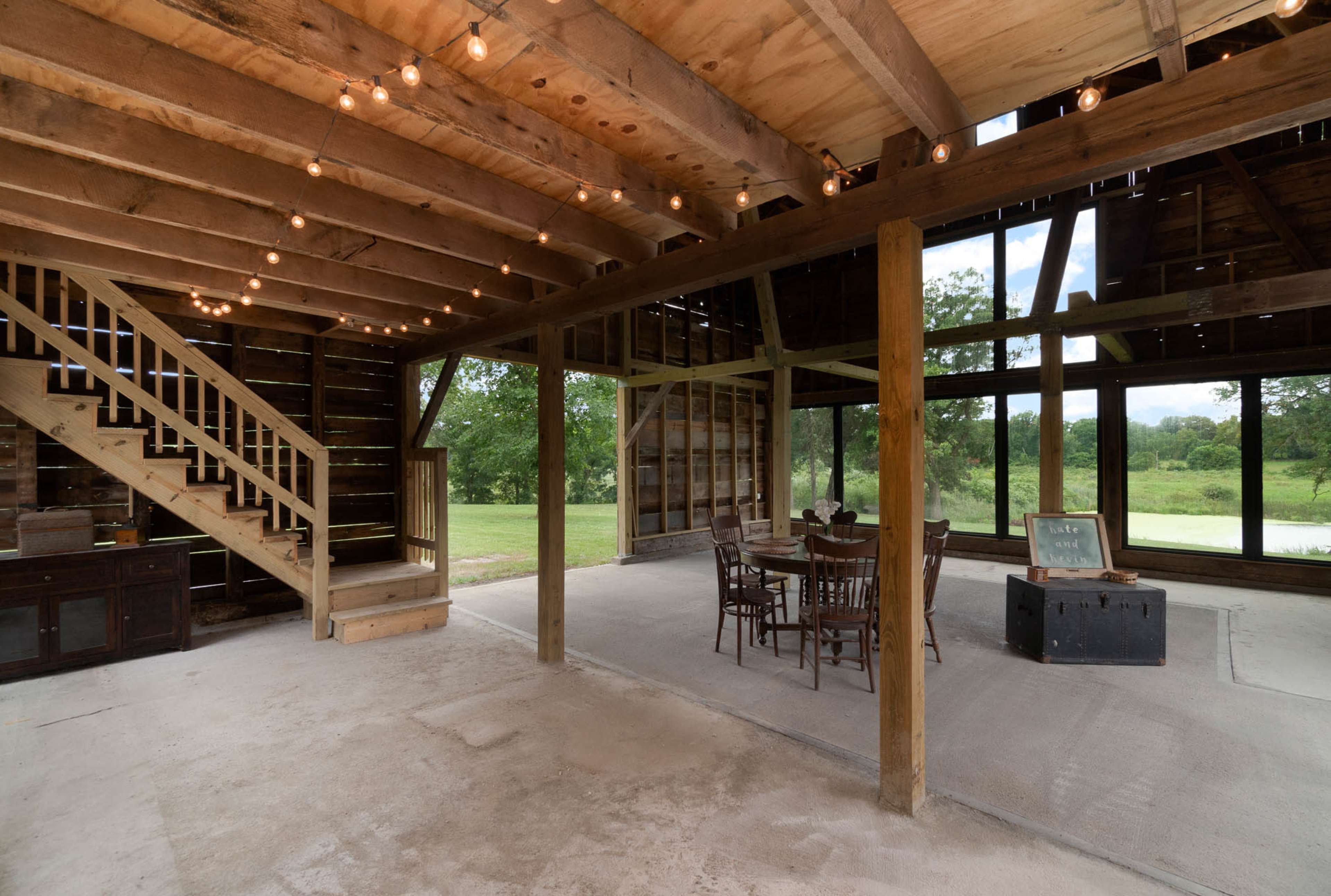The interior of a rustic, open space features wooden beams, a staircase, a dining table with chairs, and large windows that overlook a green landscape.