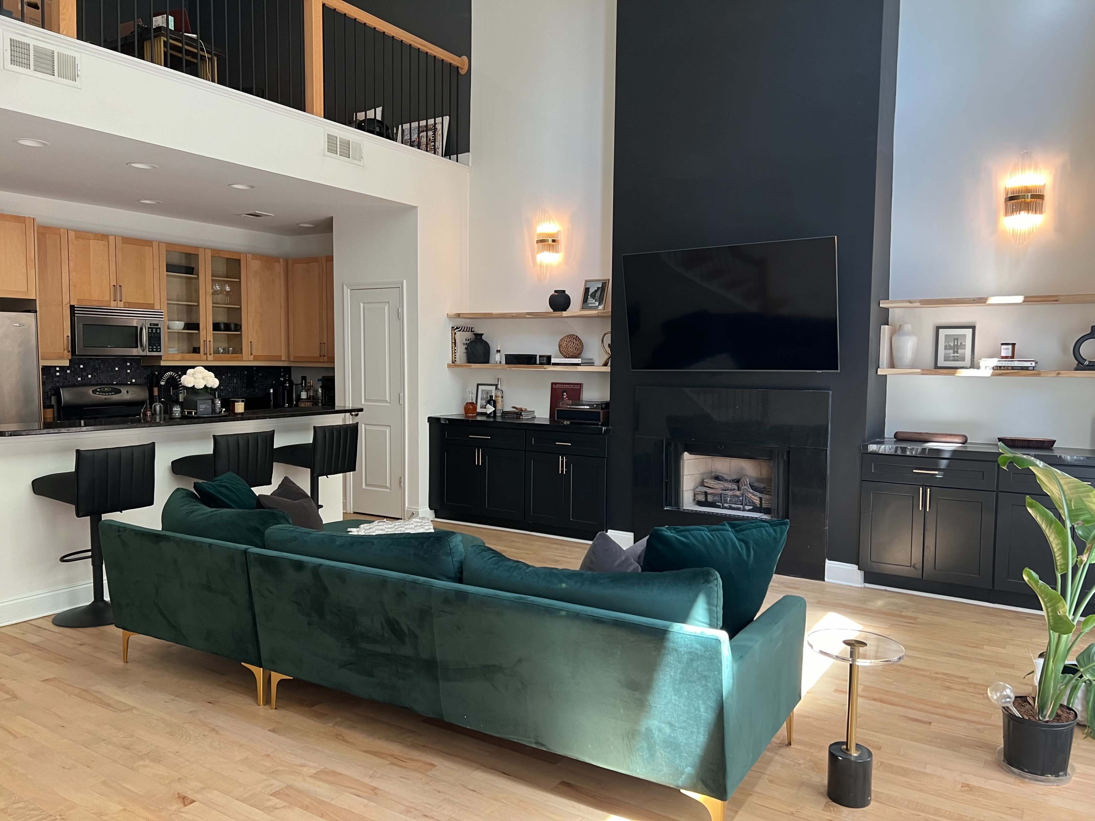 A modern living room with a green sectional sofa, a kitchen area with bar stools, and a minimalist black fireplace beneath a large flat-screen TV.