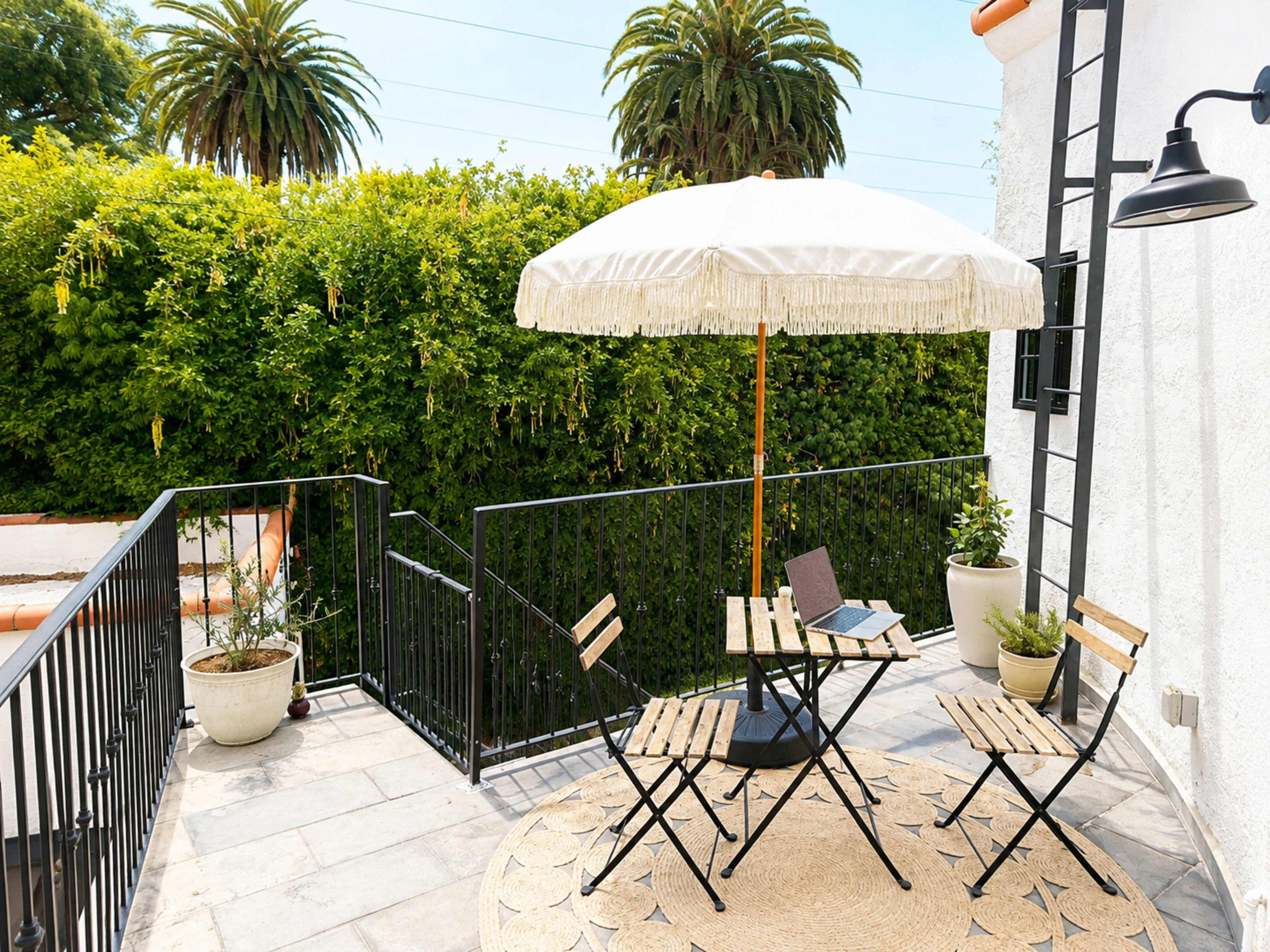 A small outdoor patio features a round table with two chairs, an umbrella overhead, and potted plants against a green hedge backdrop.