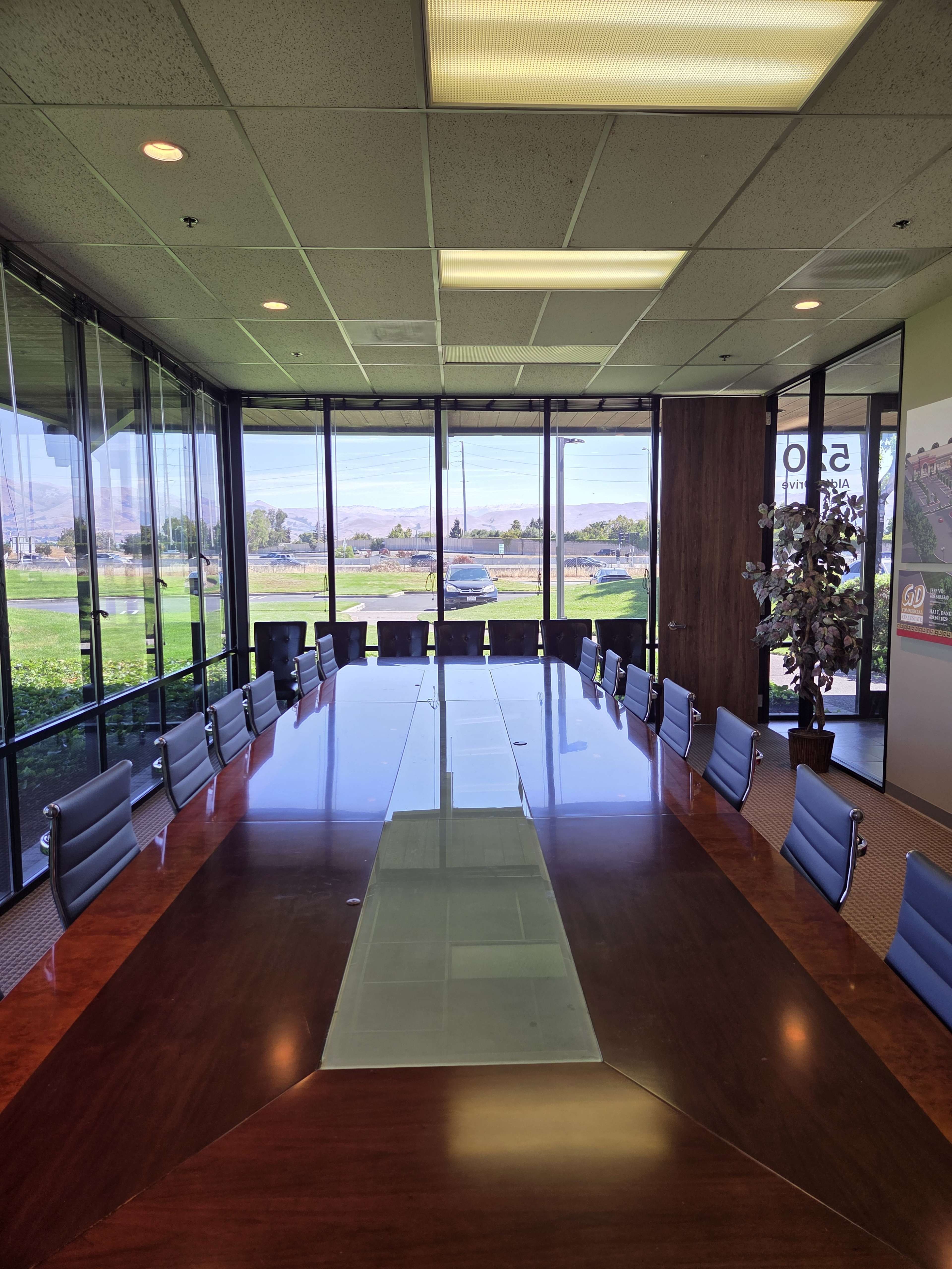A large conference room features a long wooden table surrounded by black chairs, with large windows allowing natural light and a view of the outside.