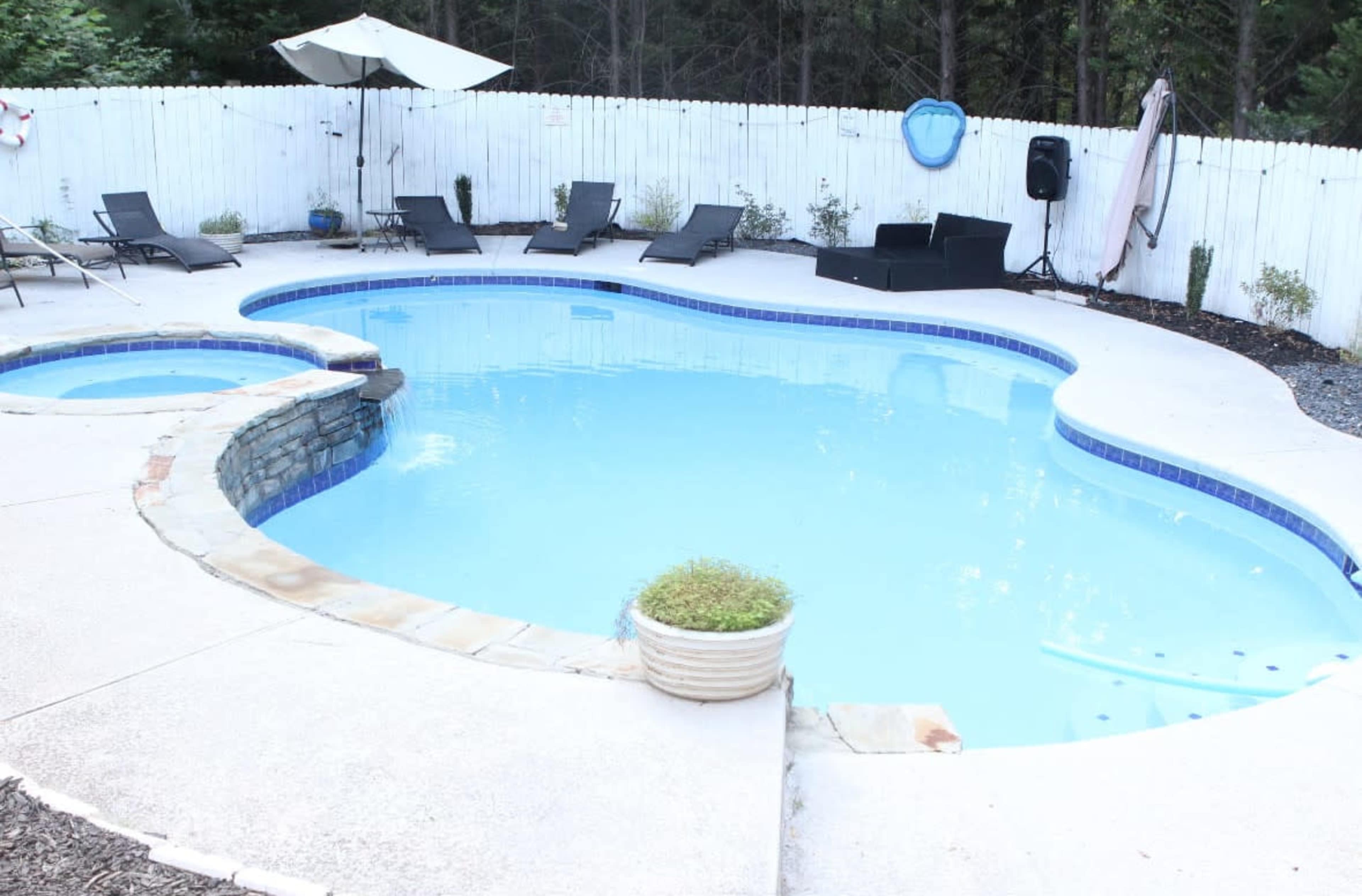 The image shows a backyard pool with a curved shape, surrounded by several lounge chairs and a patio umbrella, enclosed by a white fence and bordered by greenery.