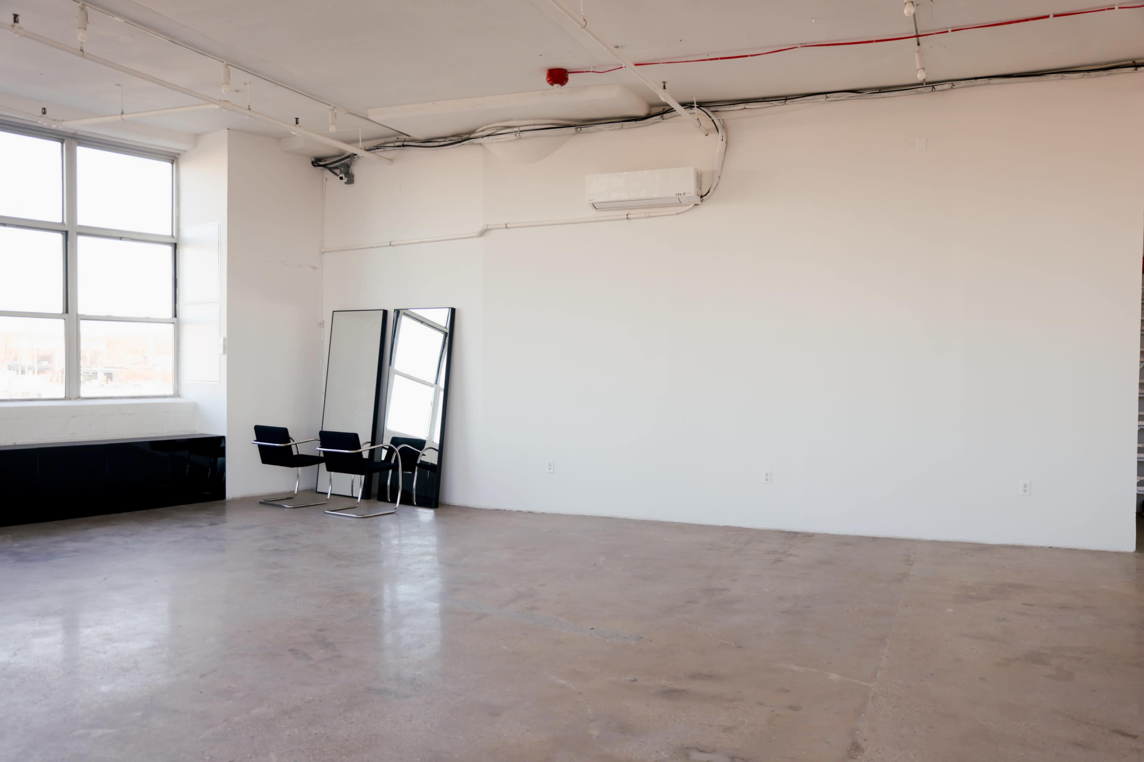 An empty, well-lit room features a large window, a mirror, and two black chairs against one wall.