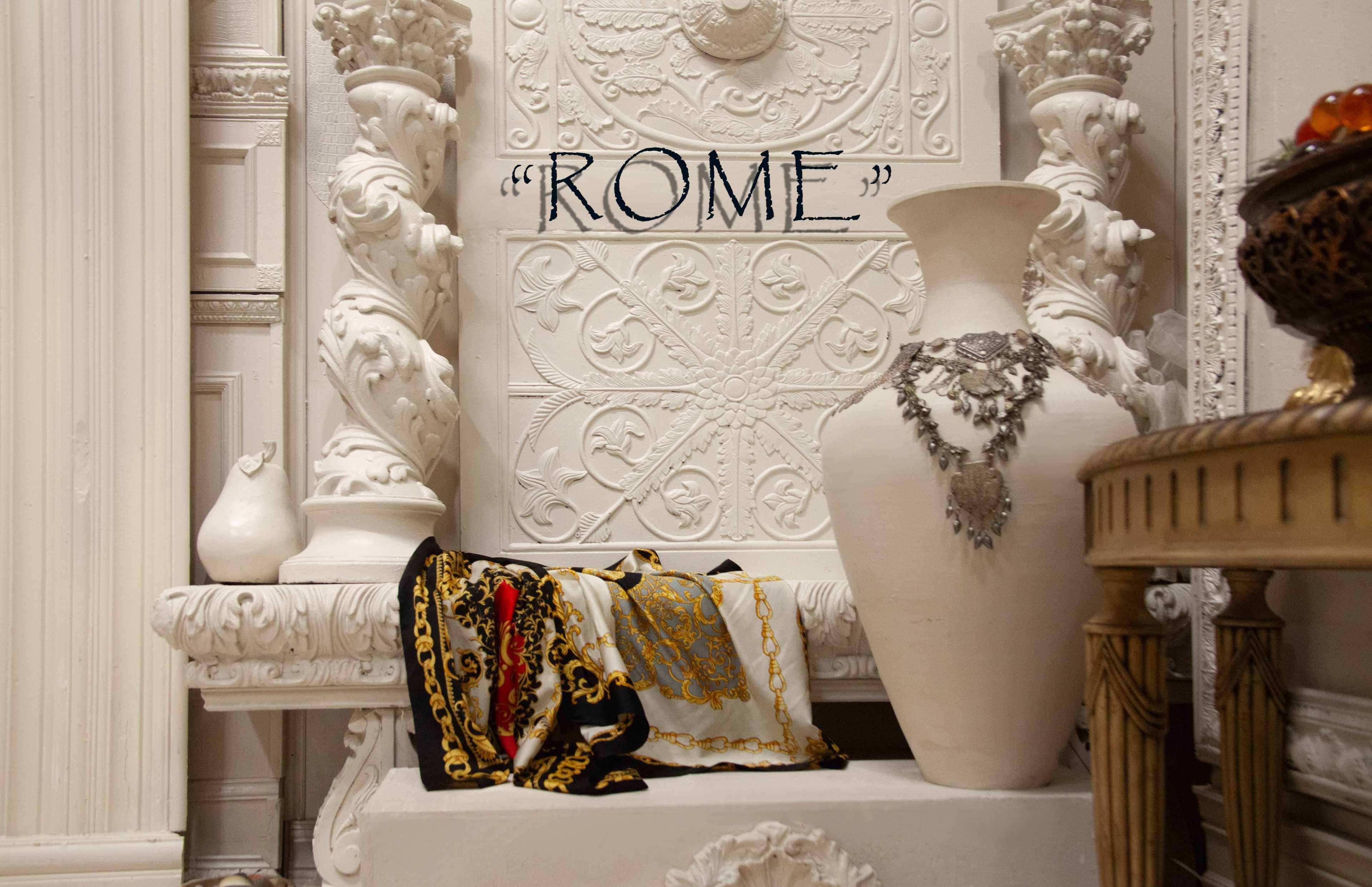 The image features an ornate white wall with decorative elements, a large vase, and a patterned scarf draped over a white ledge, with the word "ROME" prominently displayed above.