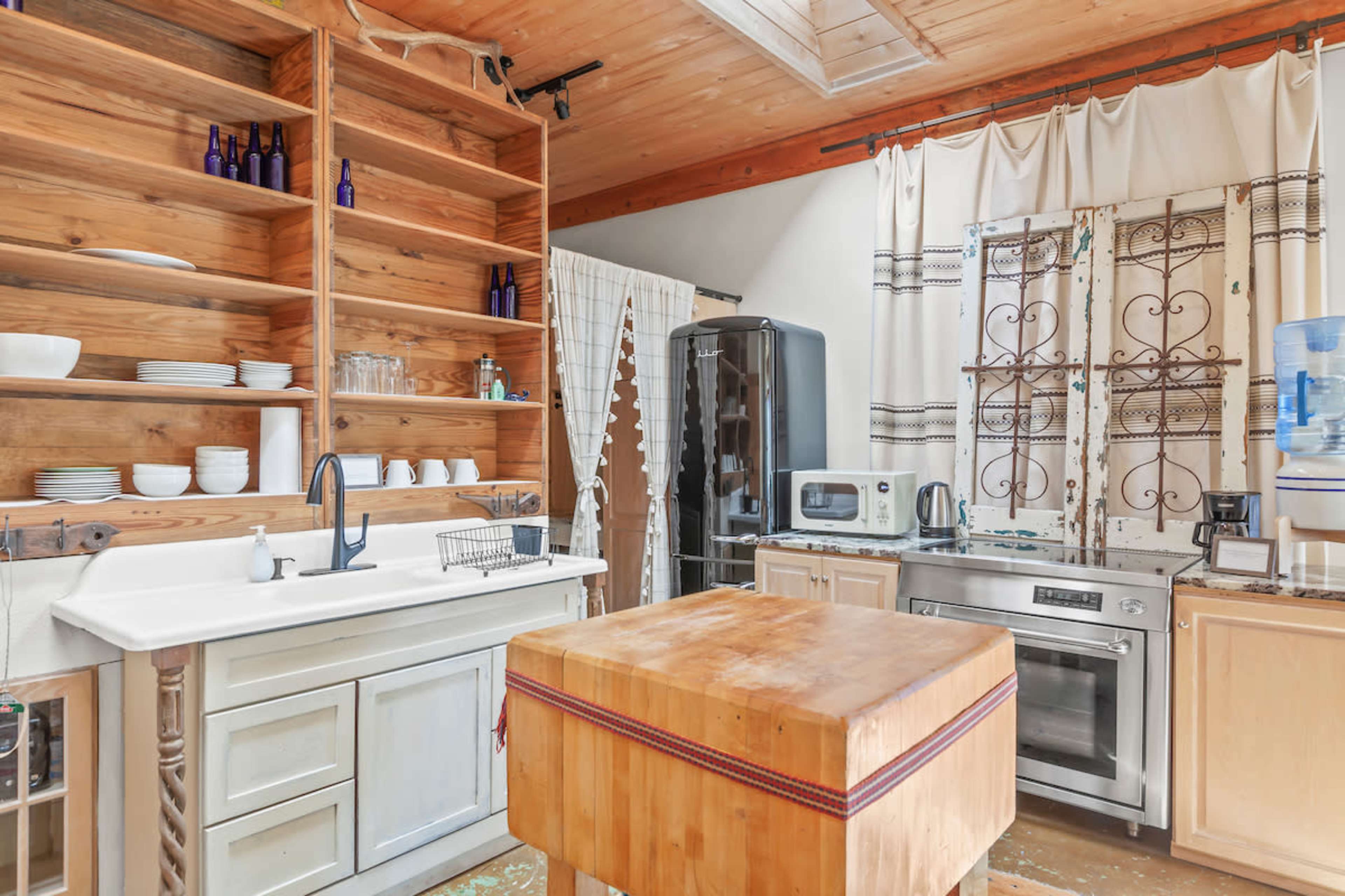 A rustic kitchen with wooden shelves, a large butcher block island, and various appliances including a refrigerator and a microwave.