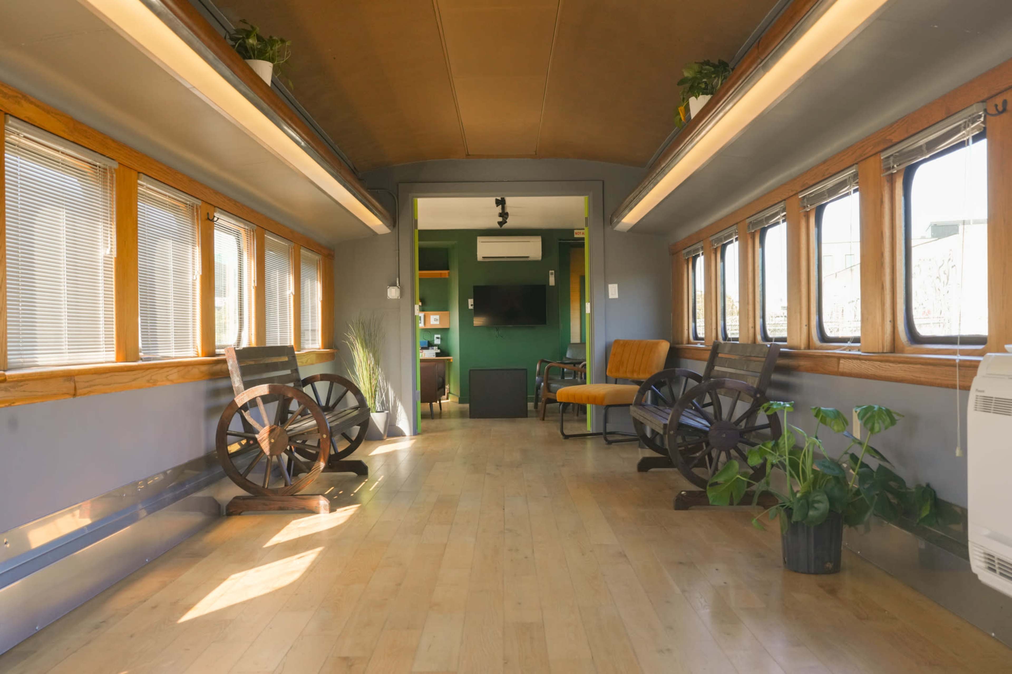 The image shows the interior of a renovated train car with wooden floors, large windows, and two decorative wheel chairs facing each other.