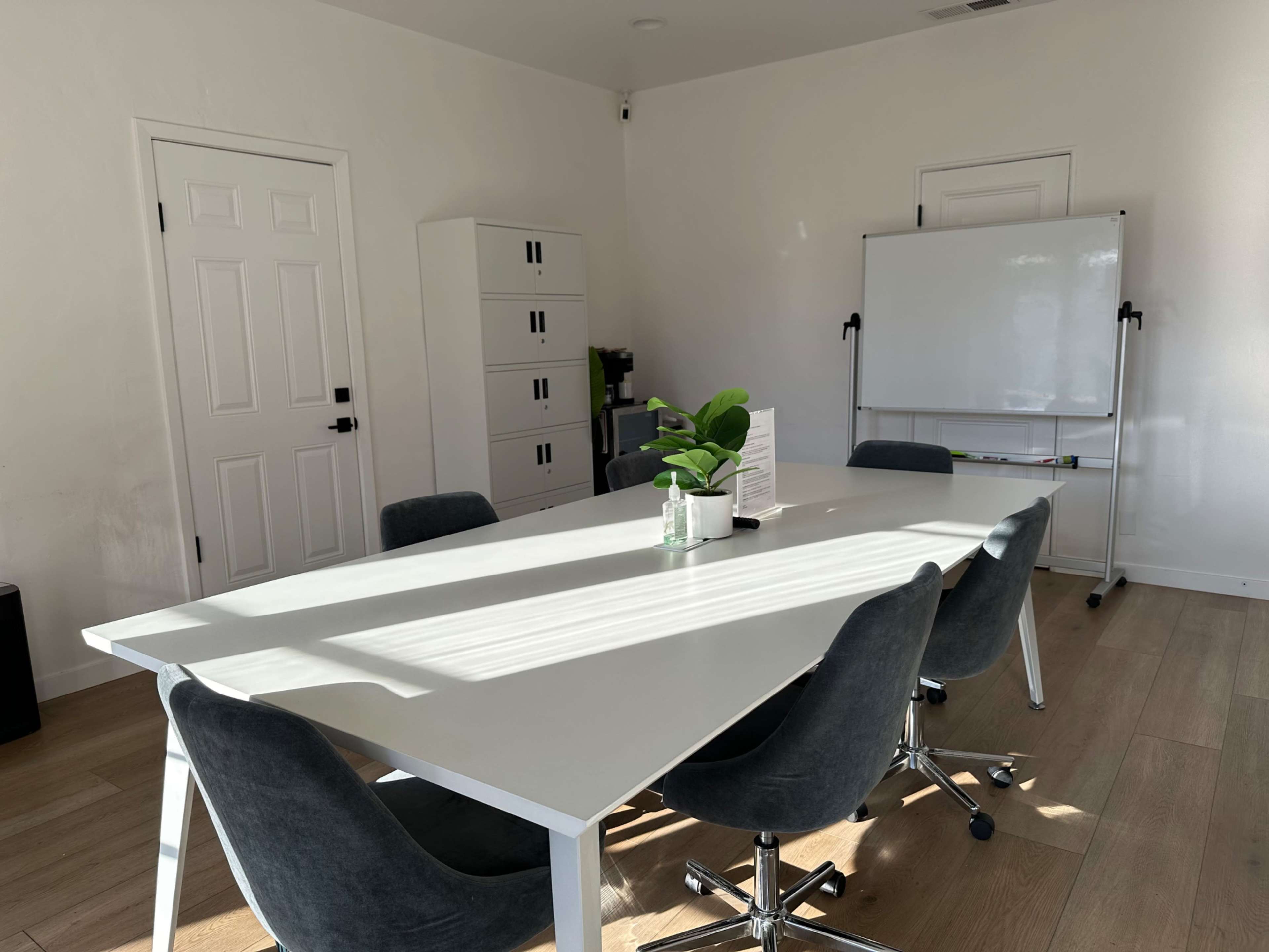 A modern conference room features a long white table surrounded by gray chairs, with a plant centerpiece and a whiteboard in the background.