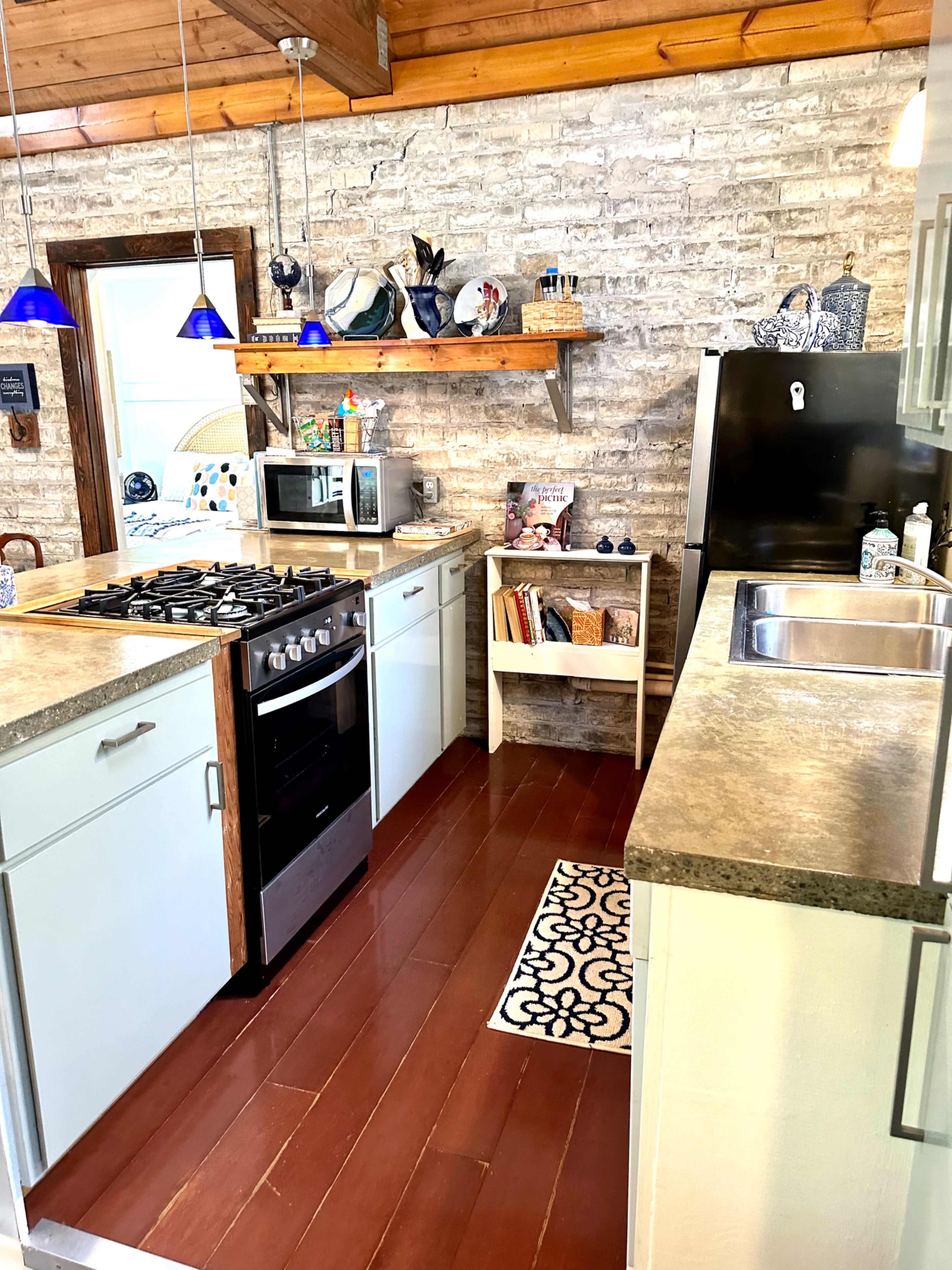 A modern kitchen with wooden ceilings, brick walls, and stainless steel appliances, featuring a gas stove, sink, and a shelf displaying decorative items.