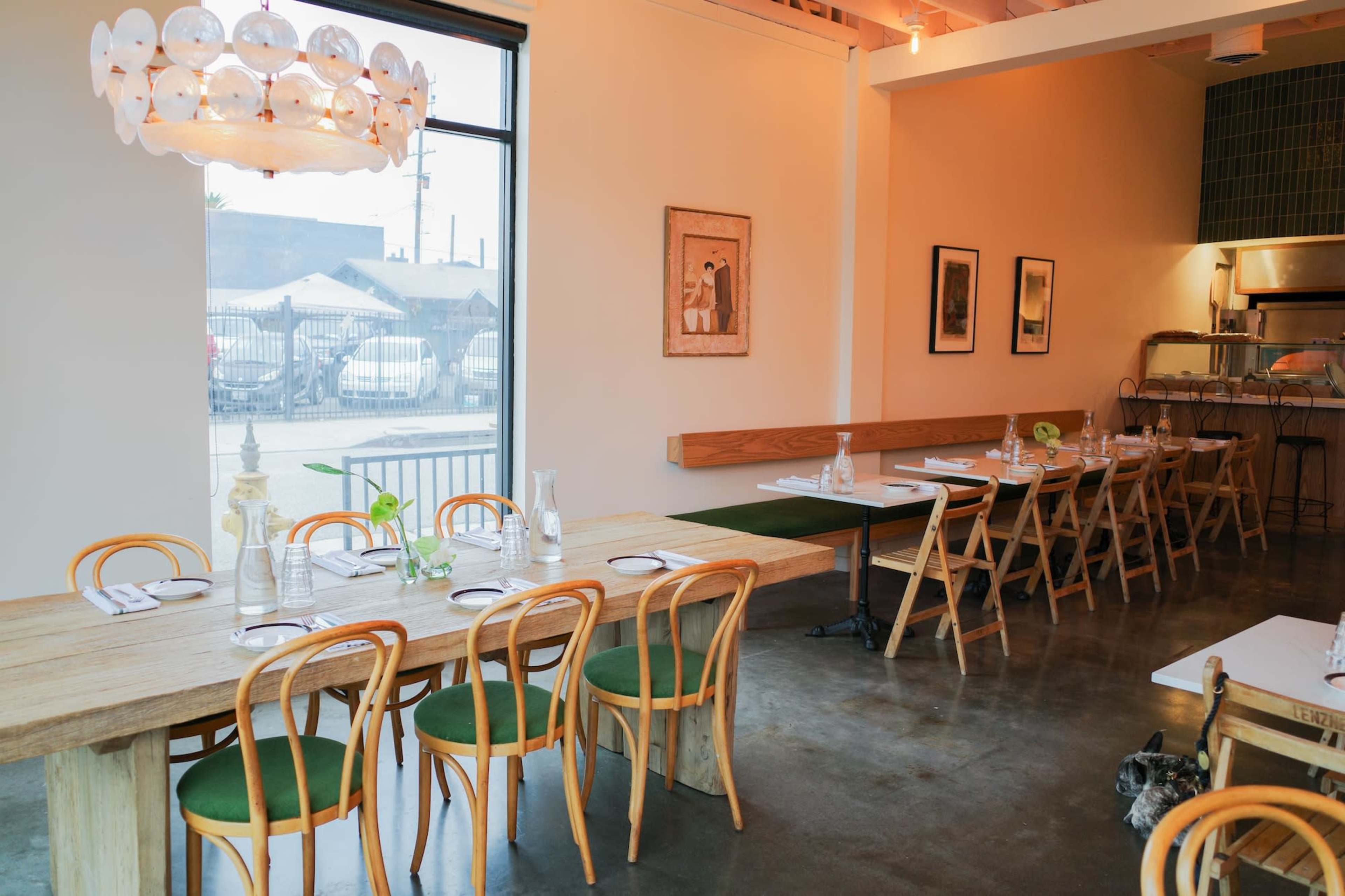 The image shows a modern restaurant interior featuring a long wooden table with green chairs, a large window, and simple decor.