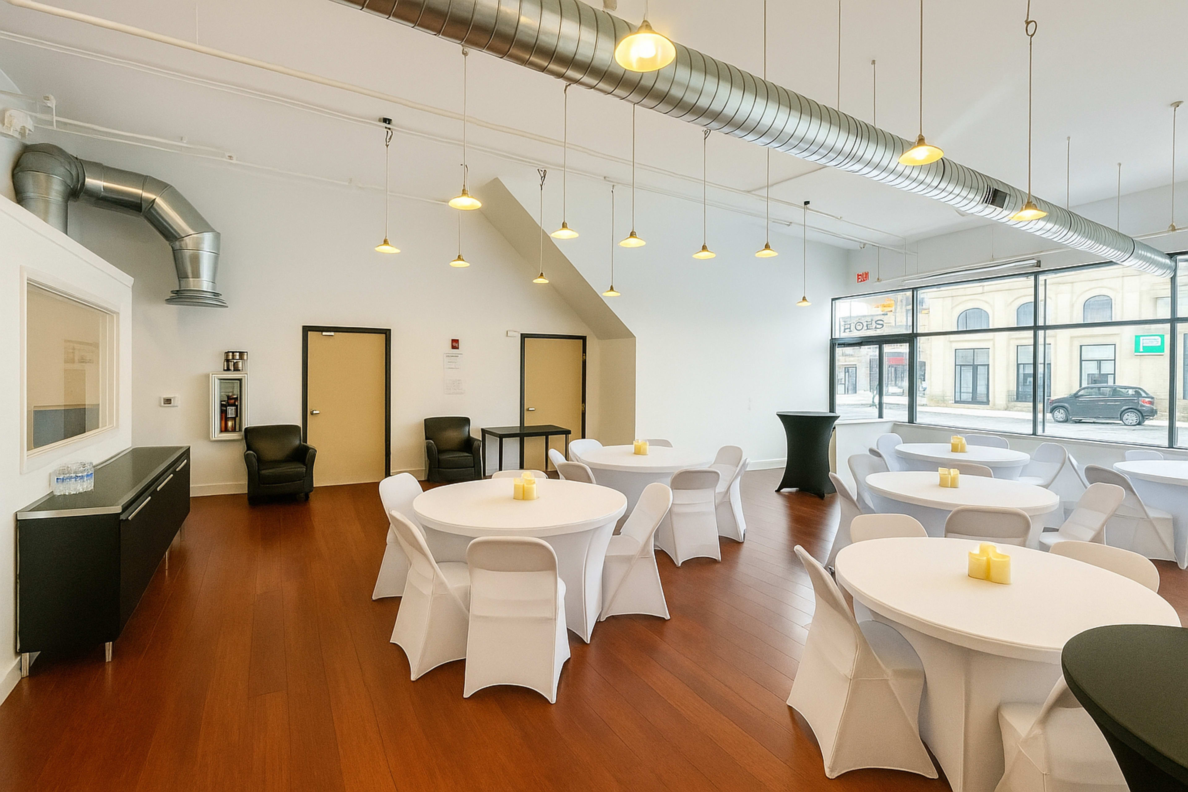 The image shows a modern event space with white tables, black chairs, and a large window letting in natural light.