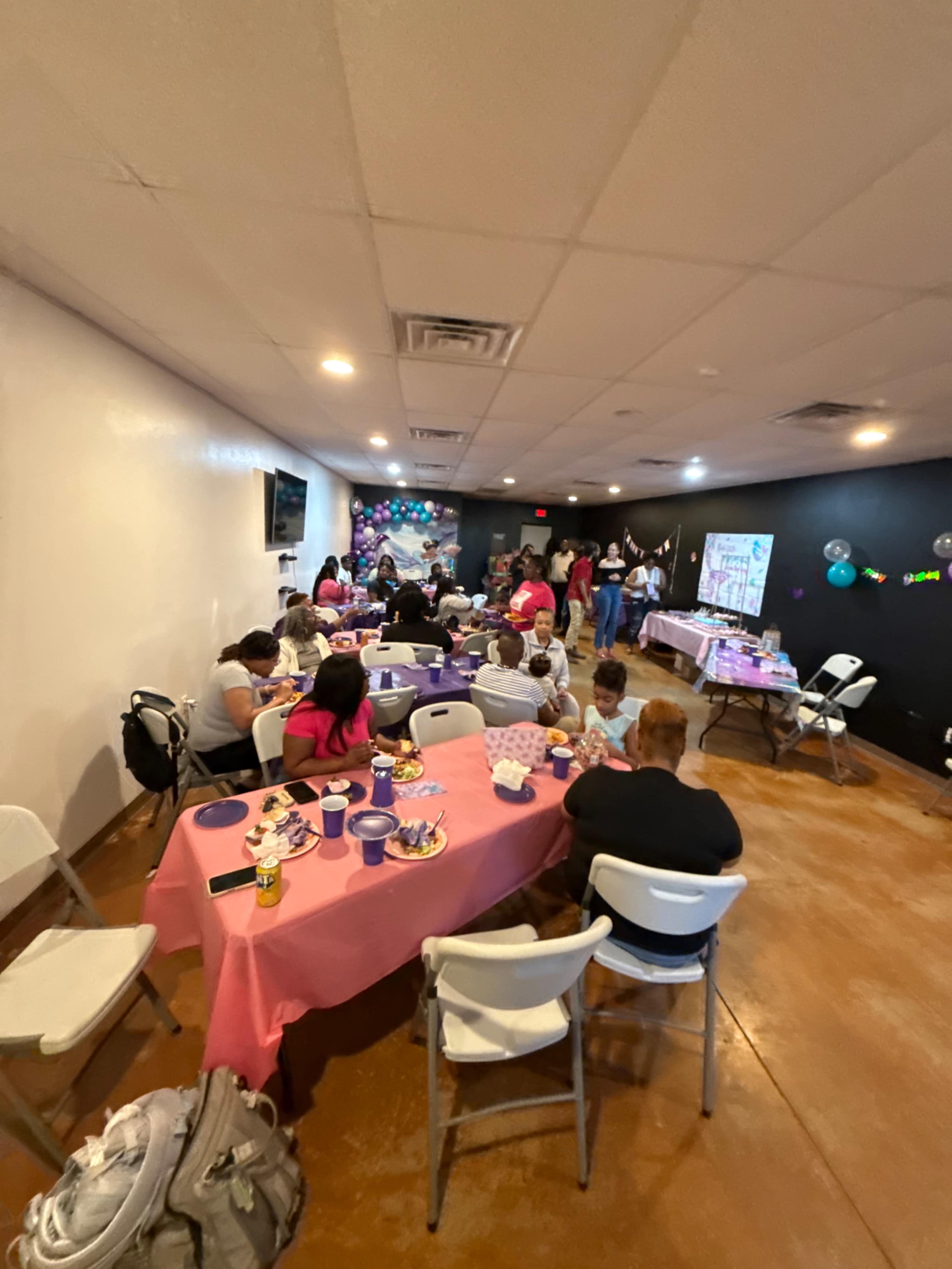 A gathering is taking place in a room with tables covered in pink and purple tablecloths, where people are sitting and enjoying food and conversations.