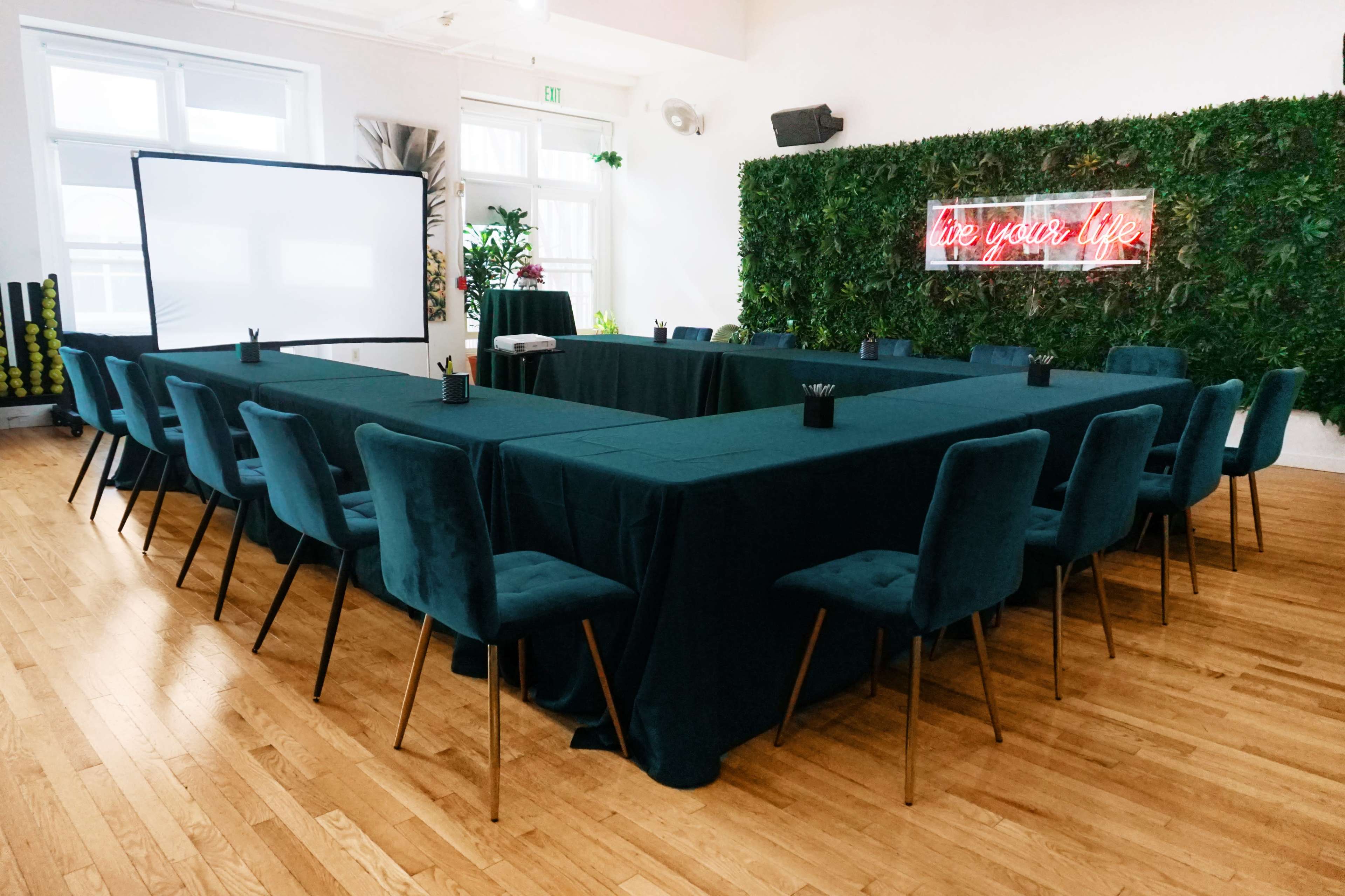 A rectangular conference room setup features a large table with teal linens surrounded by chairs, with a neon sign reading "live your life" and a wall of greenery in the background.