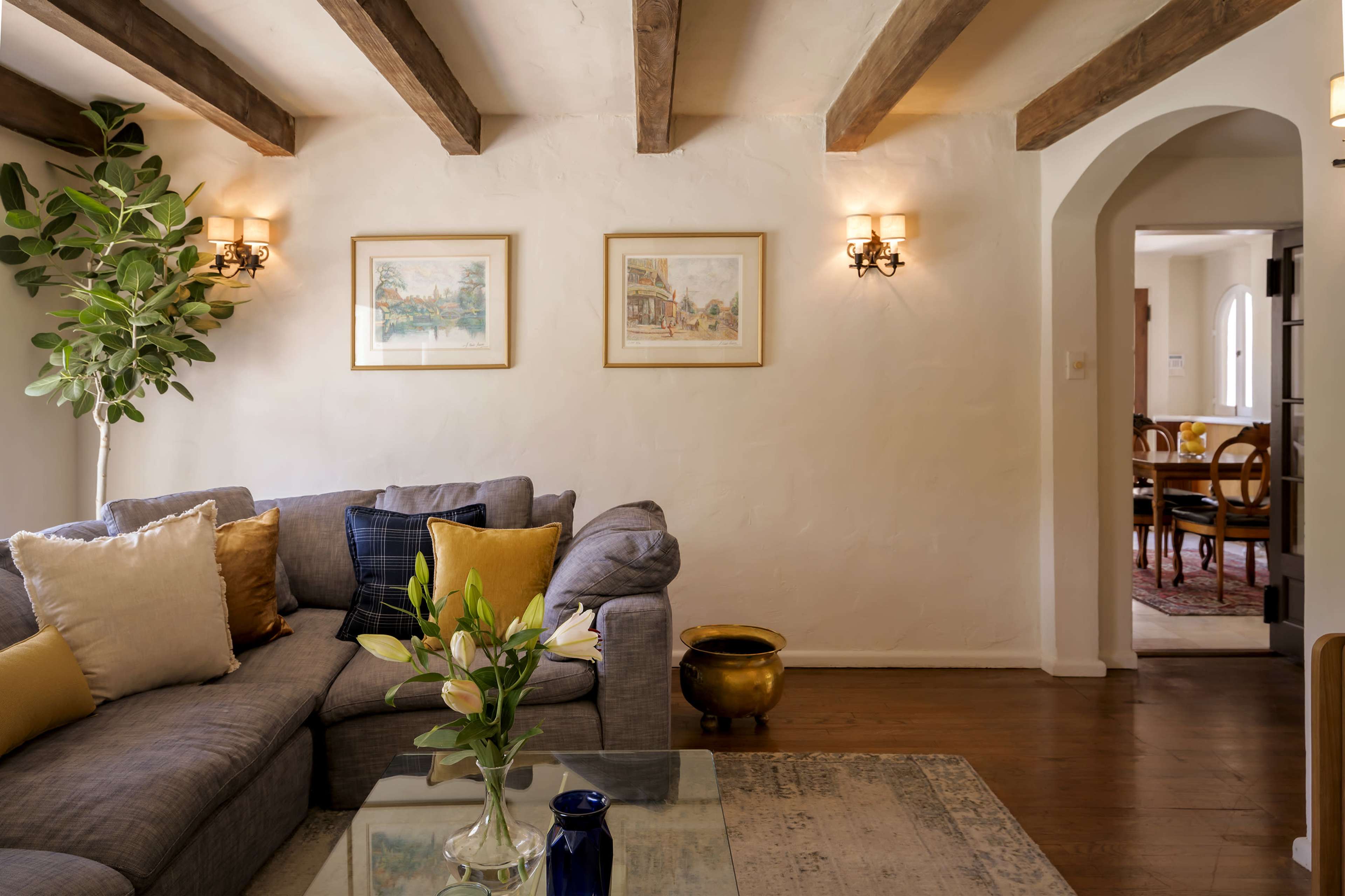 The image shows a cozy living room with a gray sectional sofa, framed artwork on the walls, and a view into a dining area through an archway.