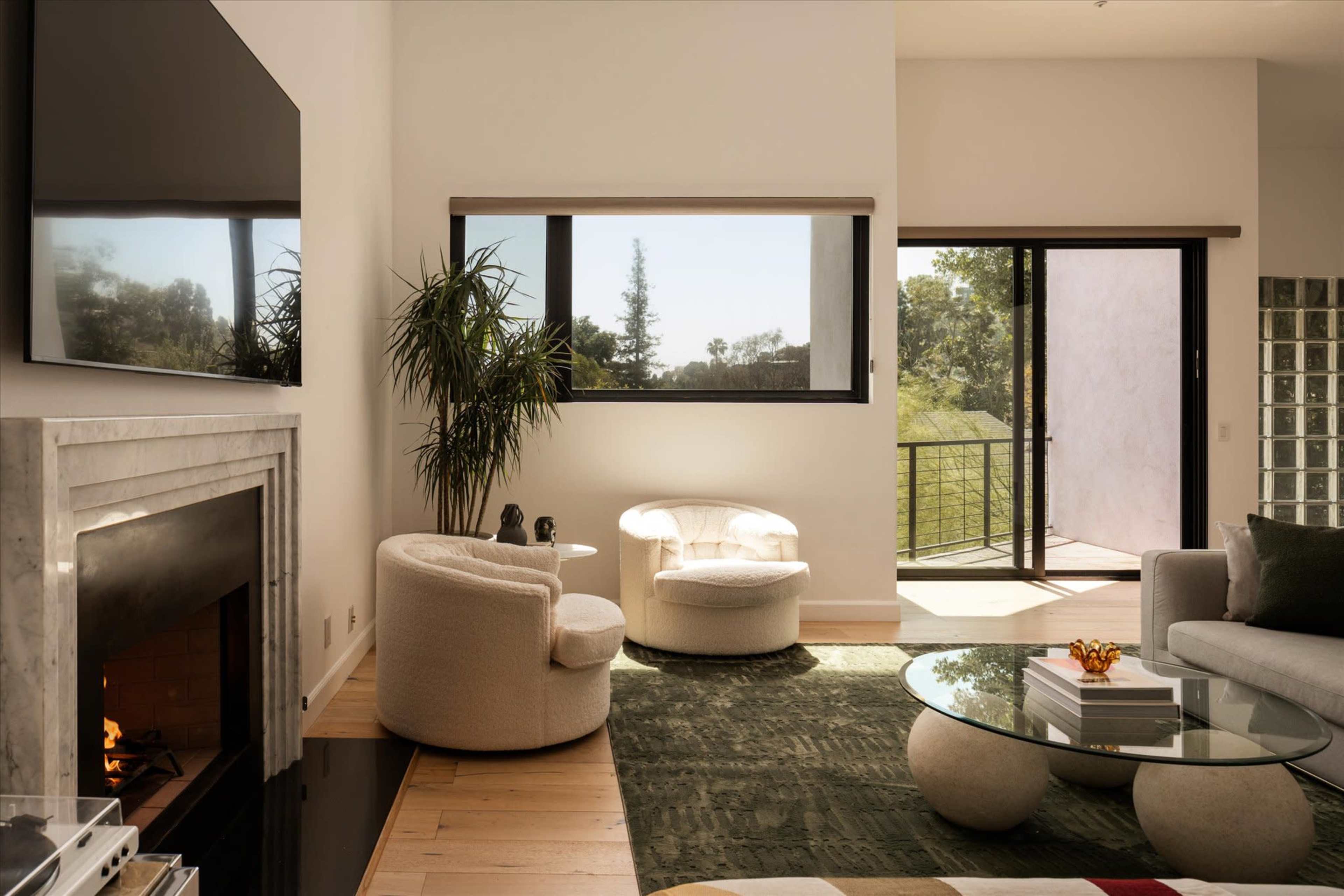 The image shows a modern living room with a fireplace, two upholstered chairs, a glass coffee table, and a large window overlooking a balcony and greenery outside.