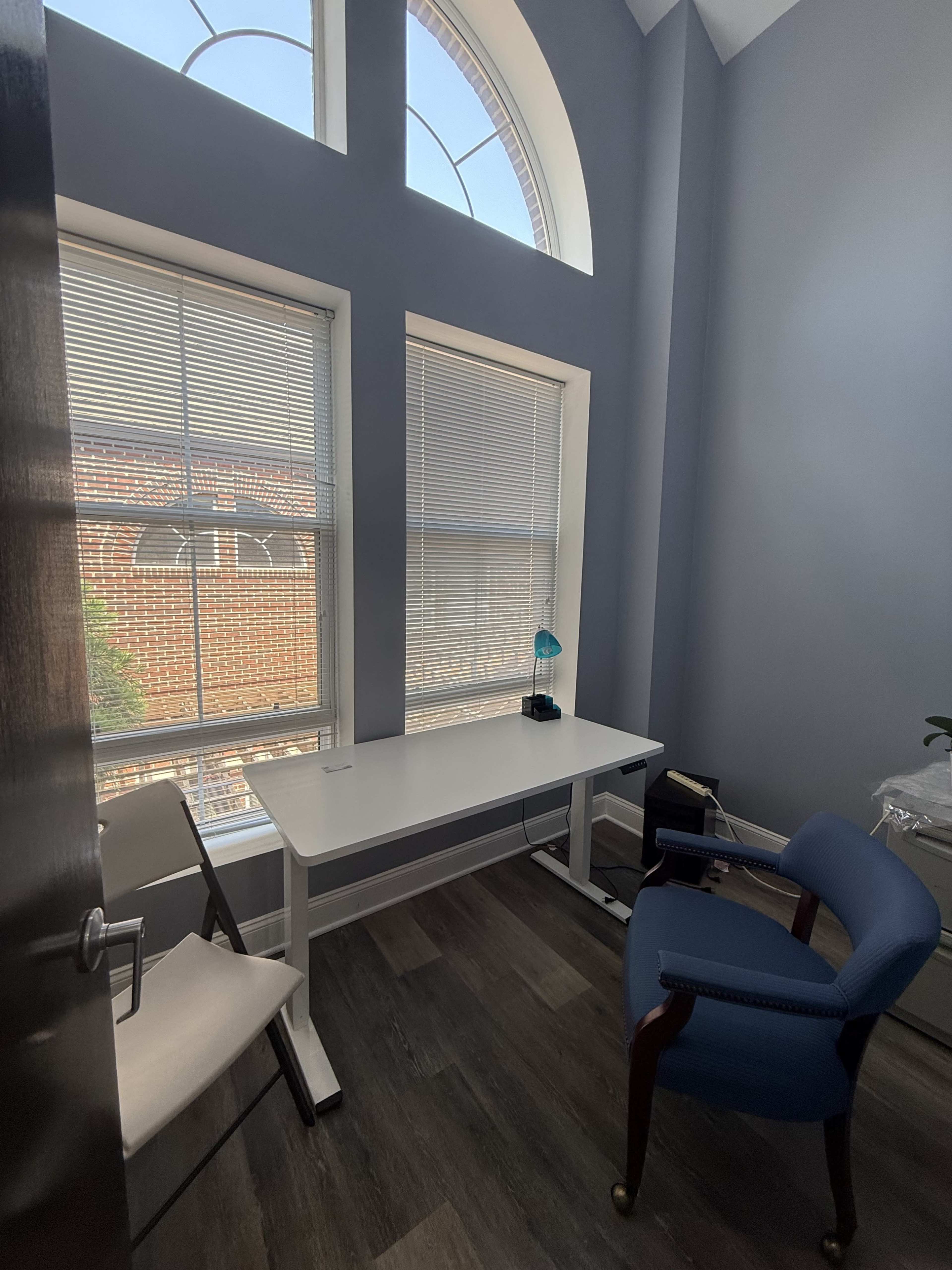The image shows a small office with a white desk, an office chair, and a folding chair beside a window with blinds.