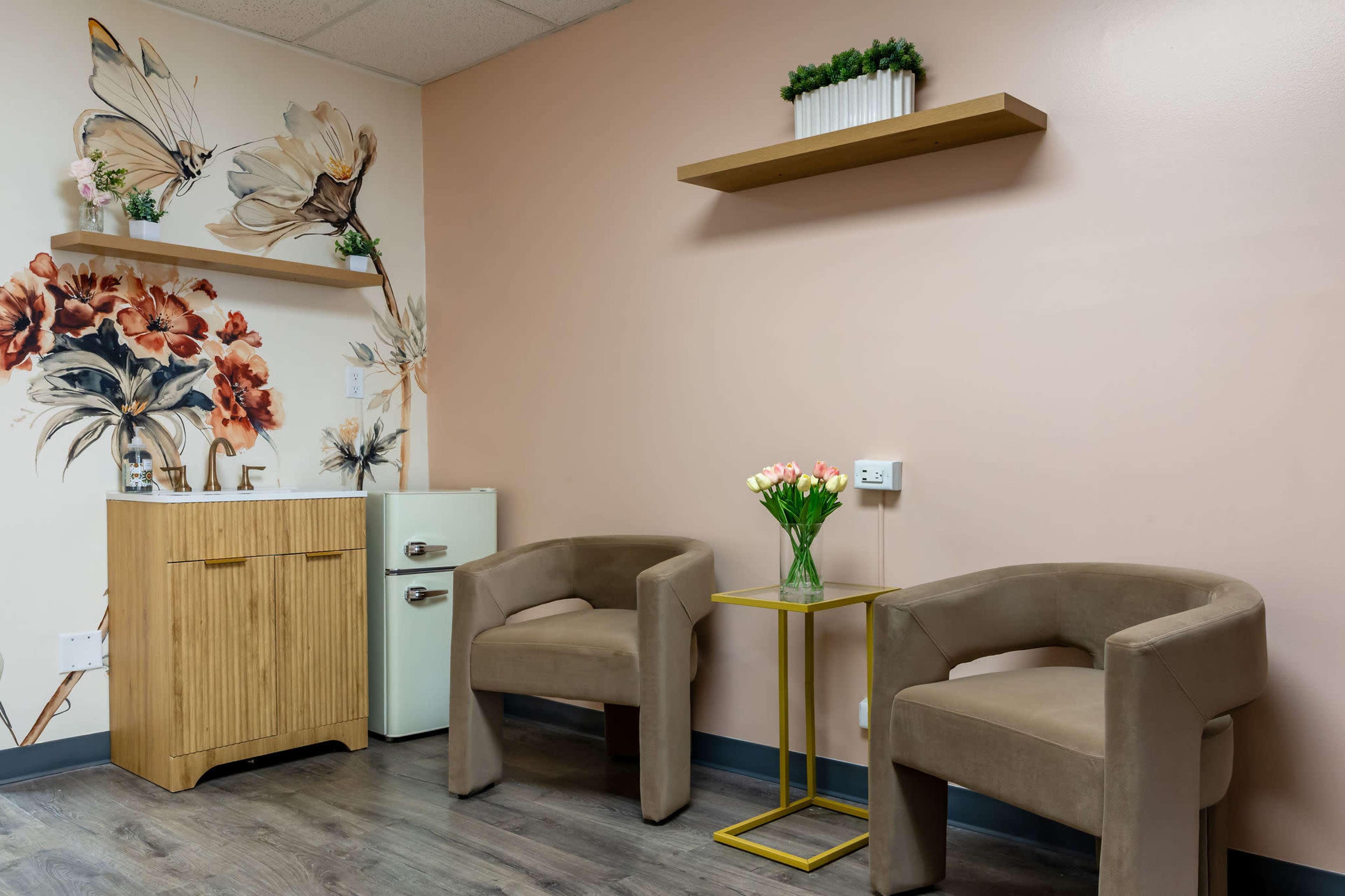 The image shows a small, elegantly designed waiting area featuring two beige armchairs, a floral wall mural, a wooden shelf, and a small table with a vase of tulips.