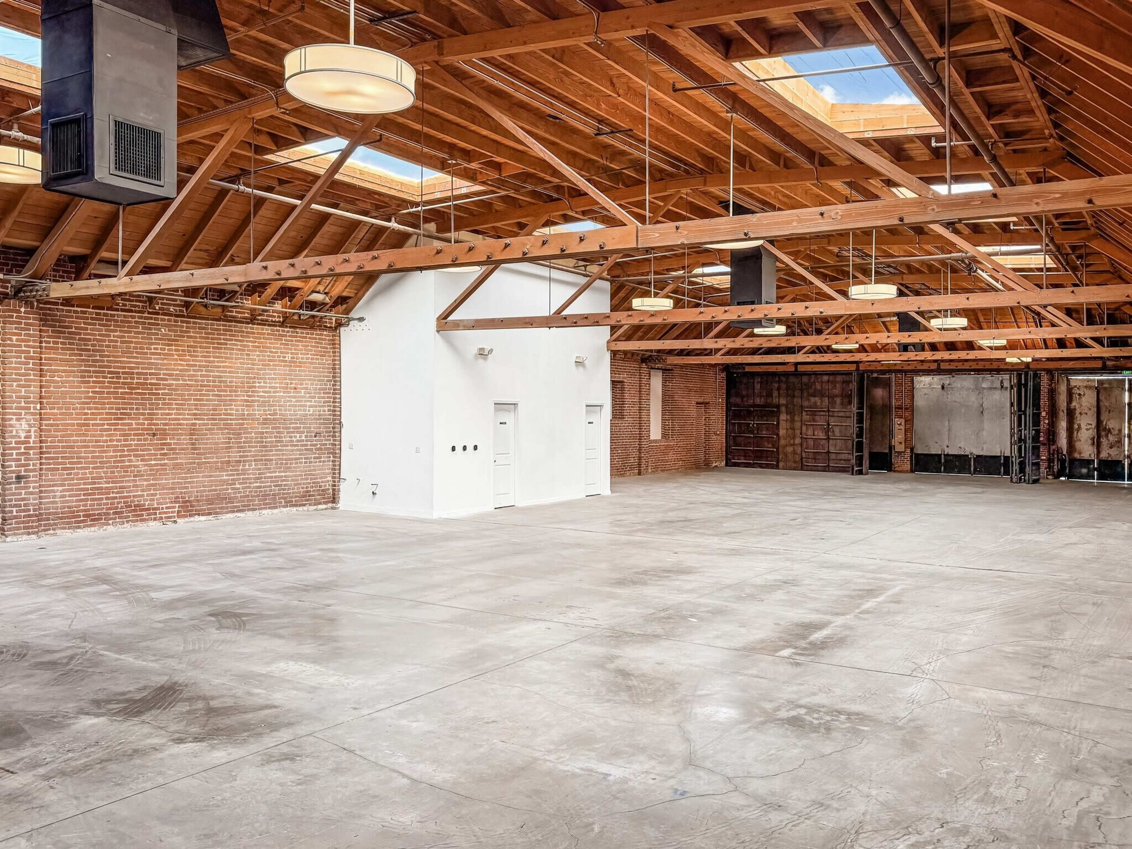 The image shows a spacious interior of a warehouse with exposed wooden beams, brick walls, and concrete flooring, featuring large skylights and minimal furnishings.