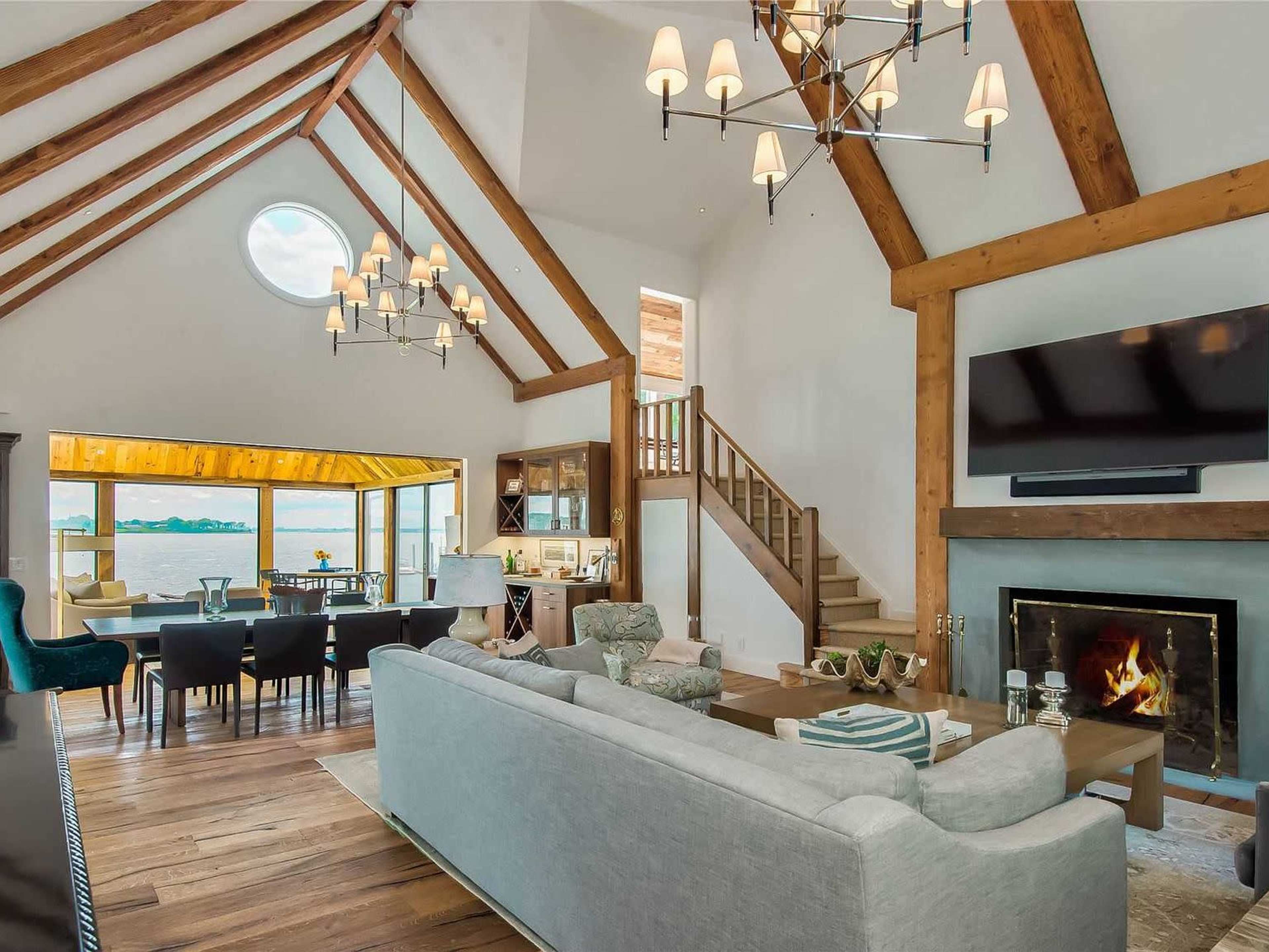 The image shows a spacious living room with high ceilings, wooden beams, a fireplace, and large windows that overlook a dining area and an outdoor view.