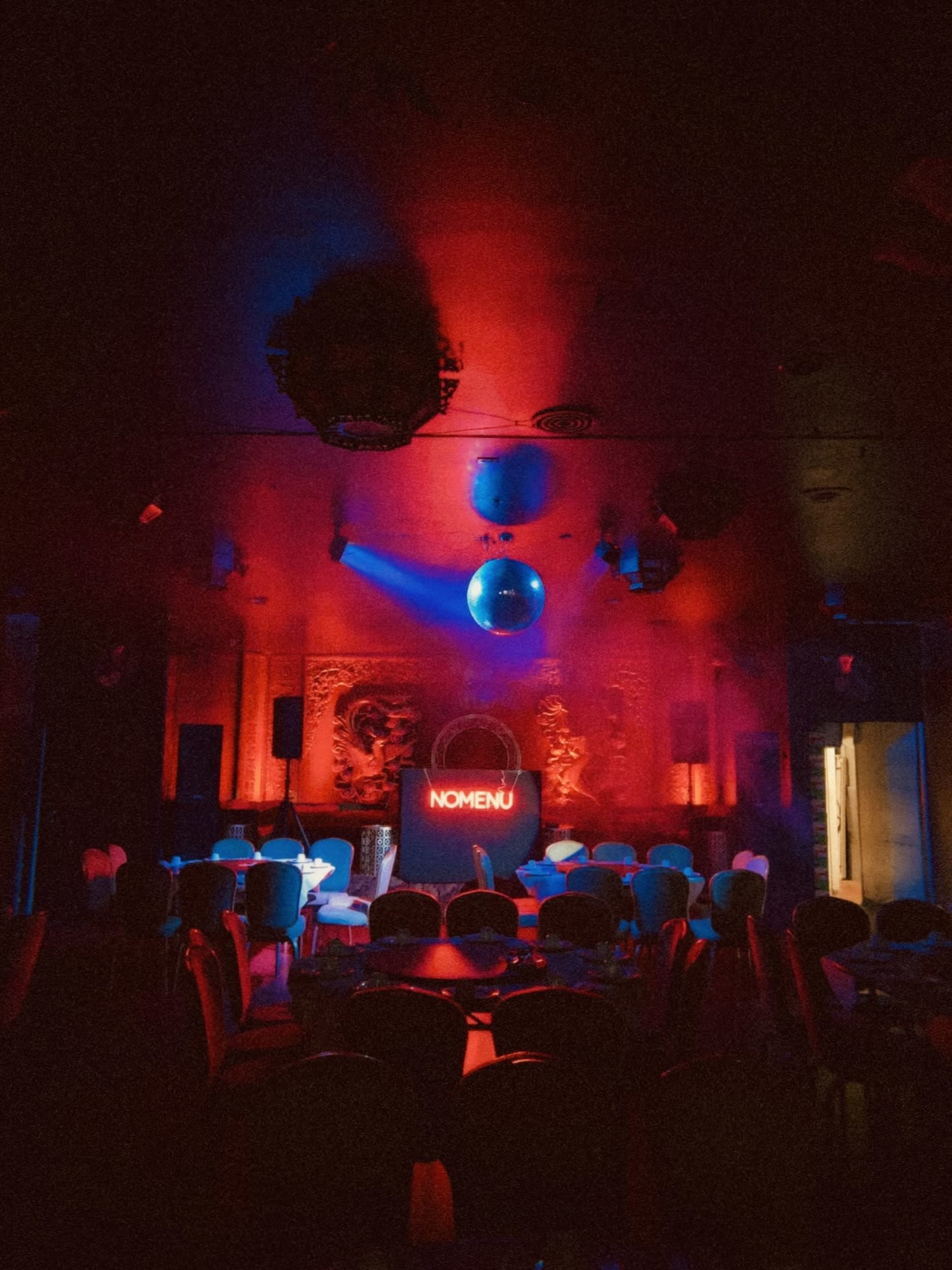 The image shows a dimly lit event space with red and blue lighting, featuring a disco ball and tables arranged in the foreground.