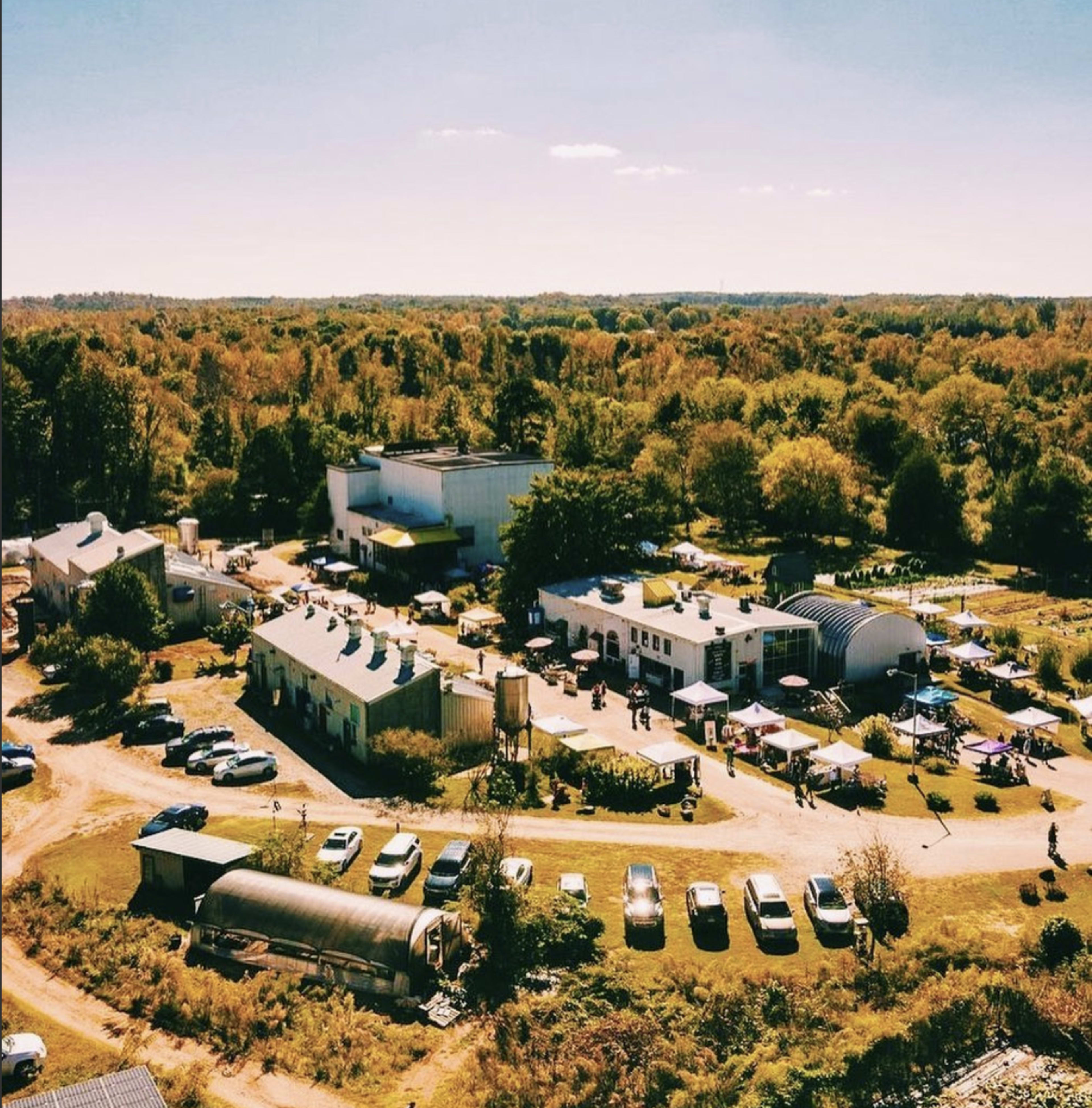 An aerial view of a farm area with several buildings, a variety of parked vehicles, and tents set up for an outdoor event surrounded by trees.