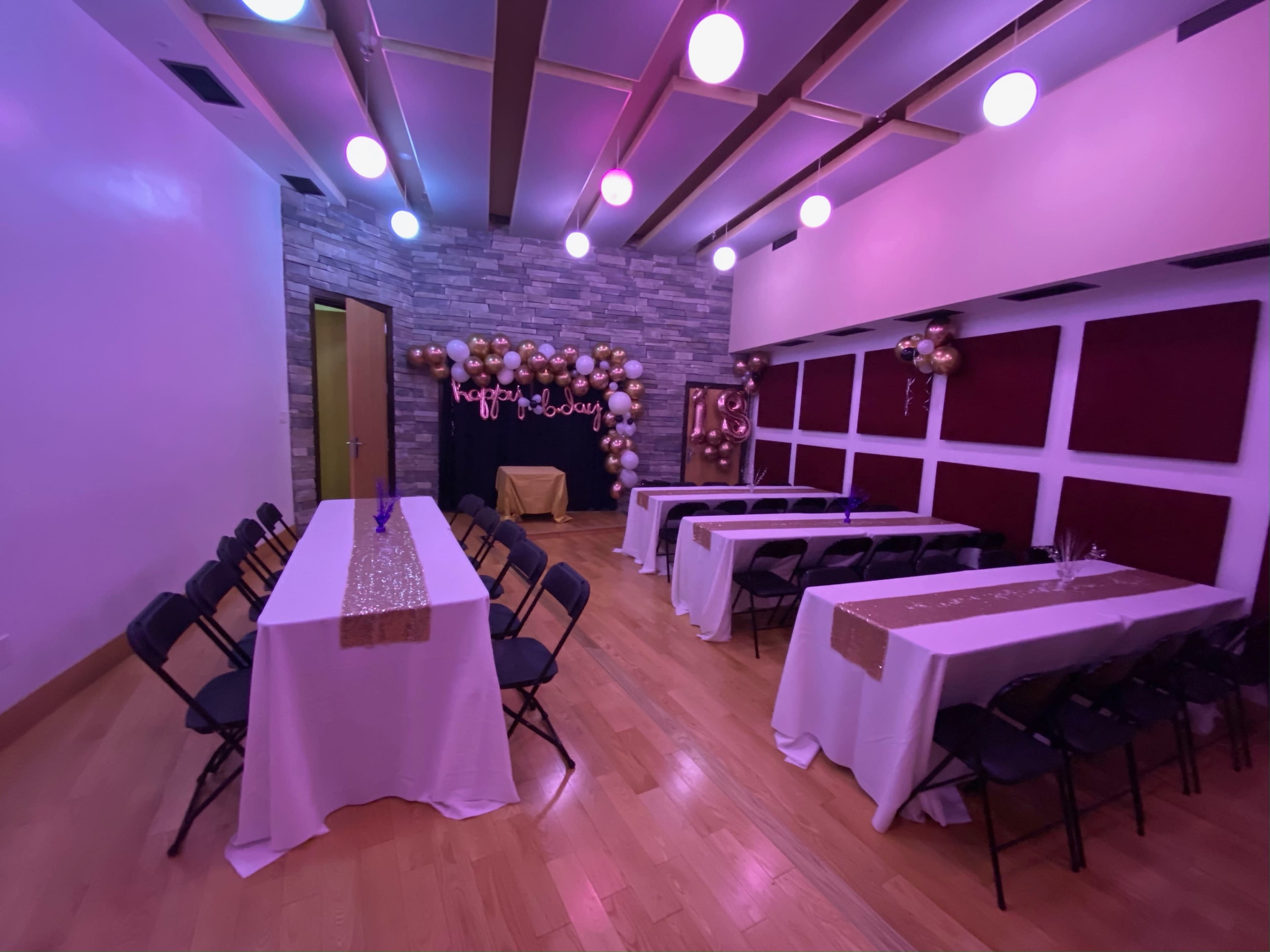 A decorated interior space set up with several tables covered in white tablecloths, balloons, and a backdrop for an event.