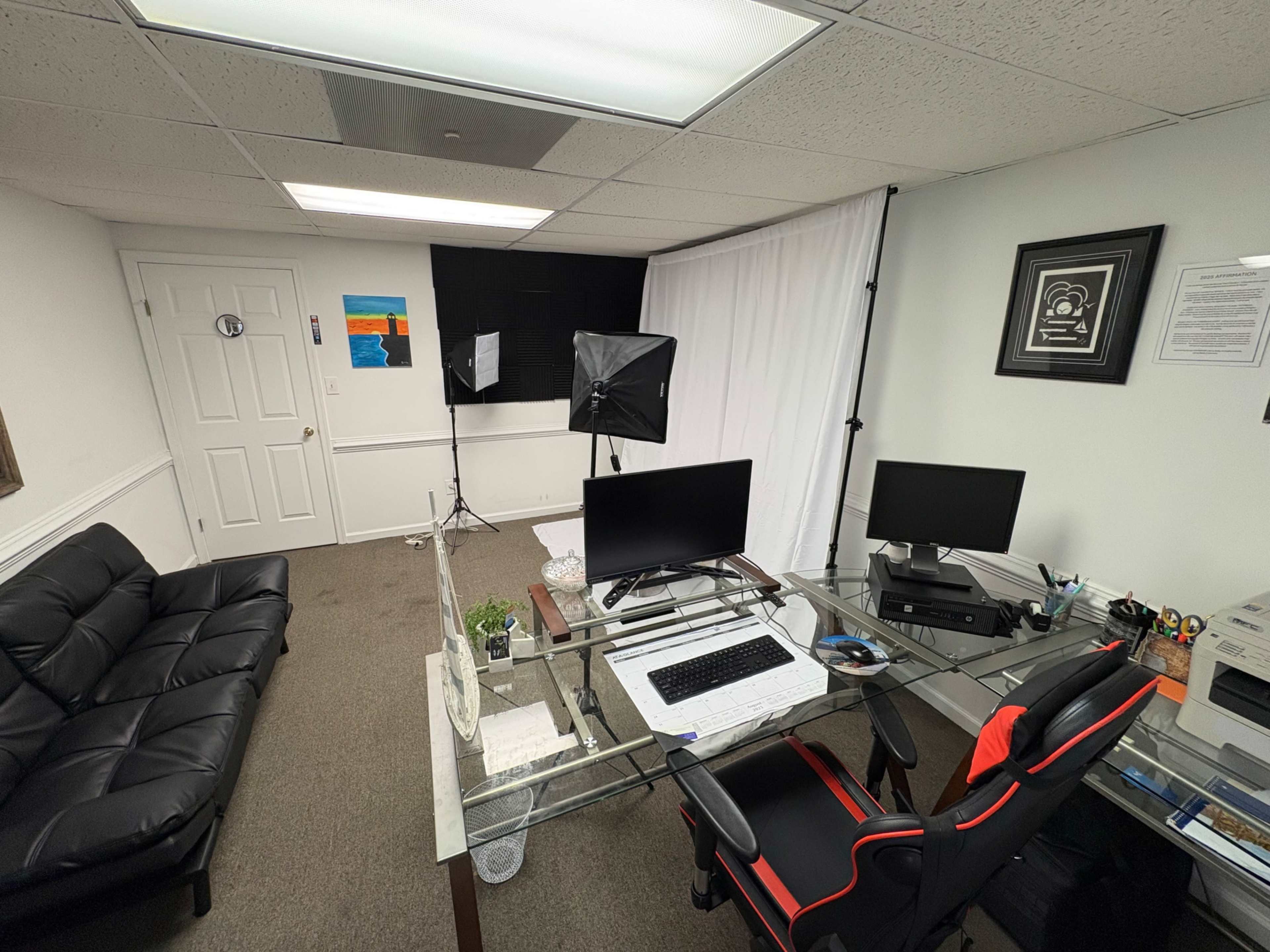 The image shows a small office space featuring a glass desk with two monitors, a black couch, and a backdrop with lighting setups for video recording.