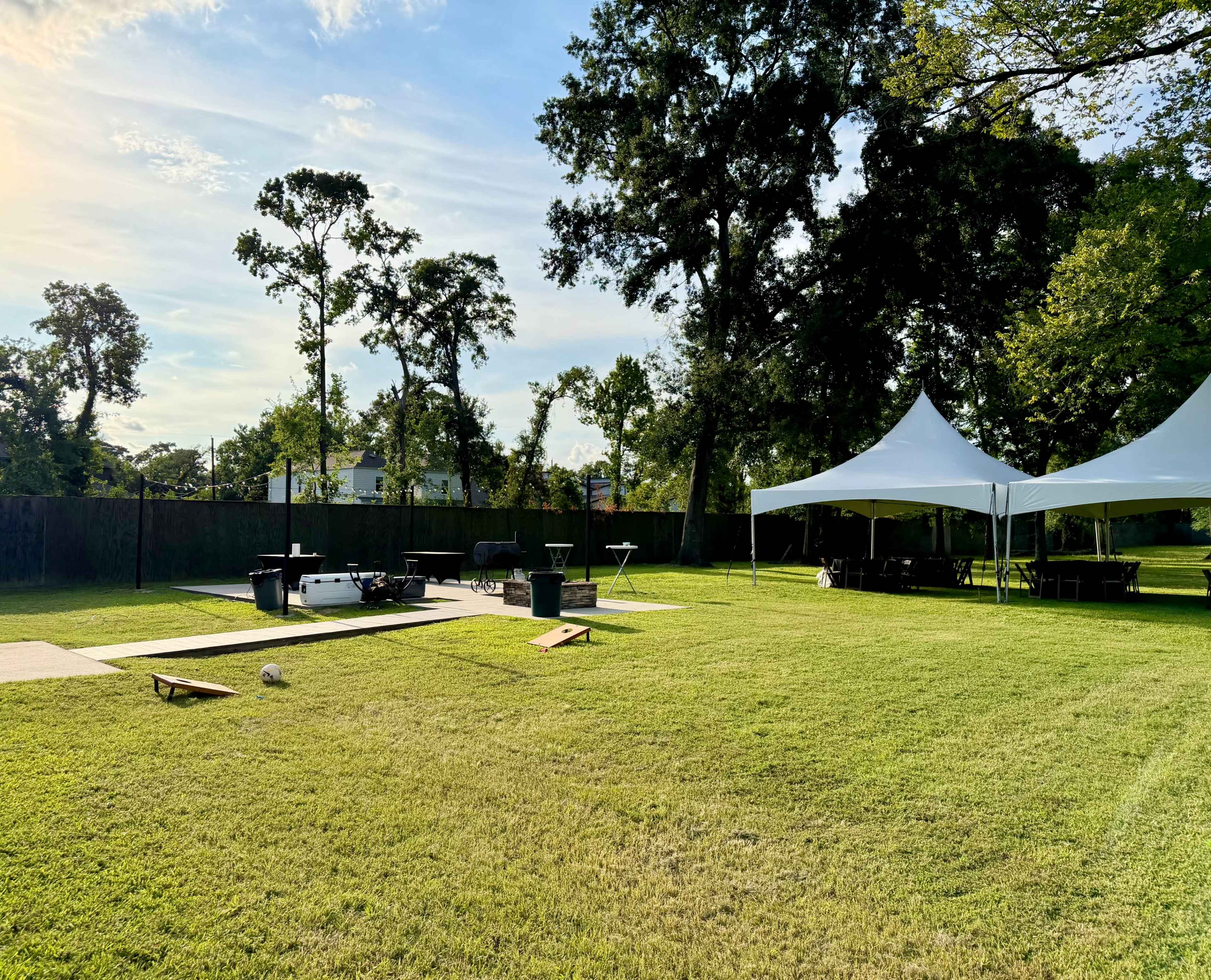 The image shows a green, open backyard featuring a white tent set up for an event, with a wooden deck, lawn games, and trees in the background.