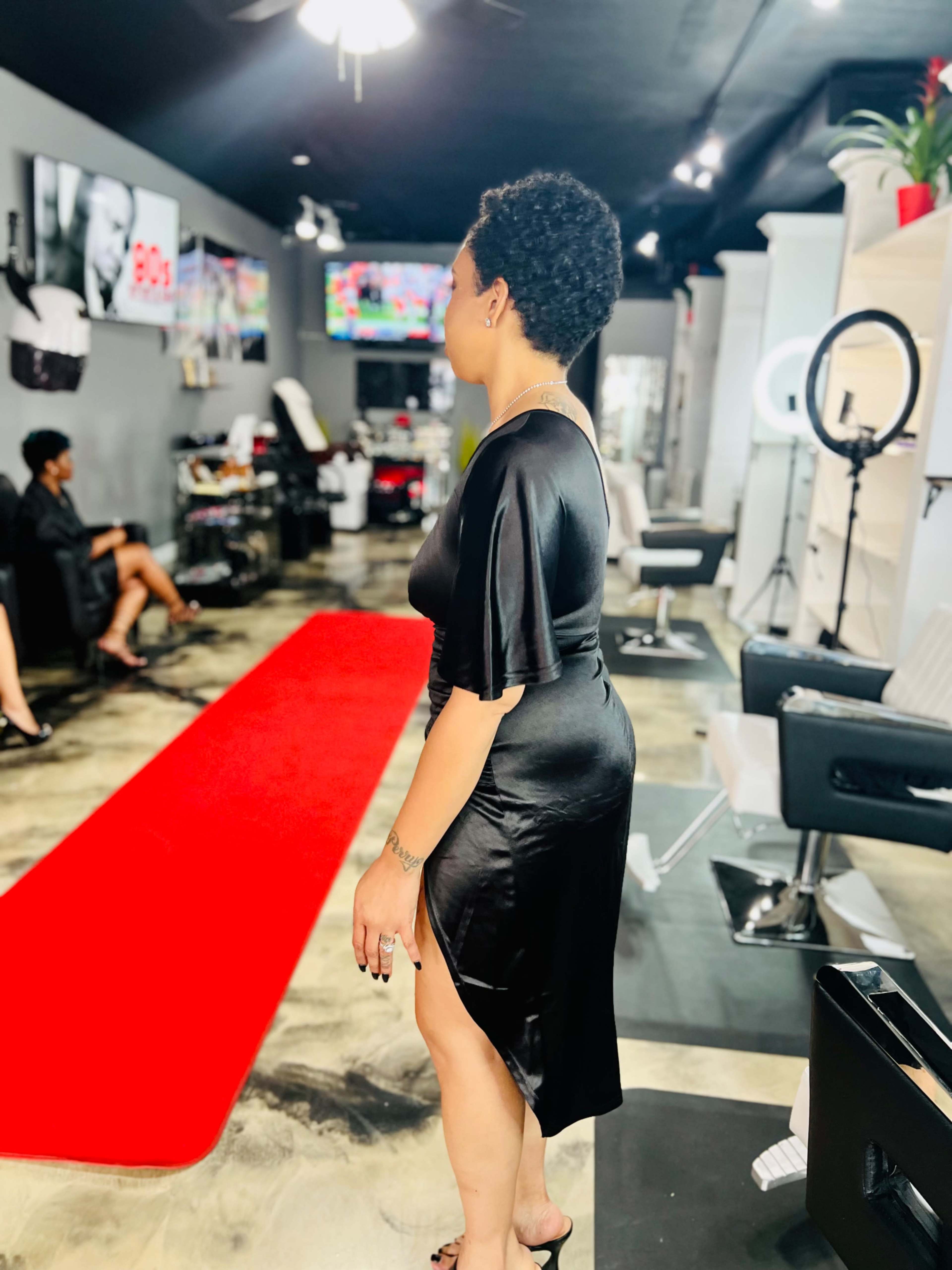 A woman in a black dress stands on a red carpet in a hair salon, while another seated woman is visible in the background.