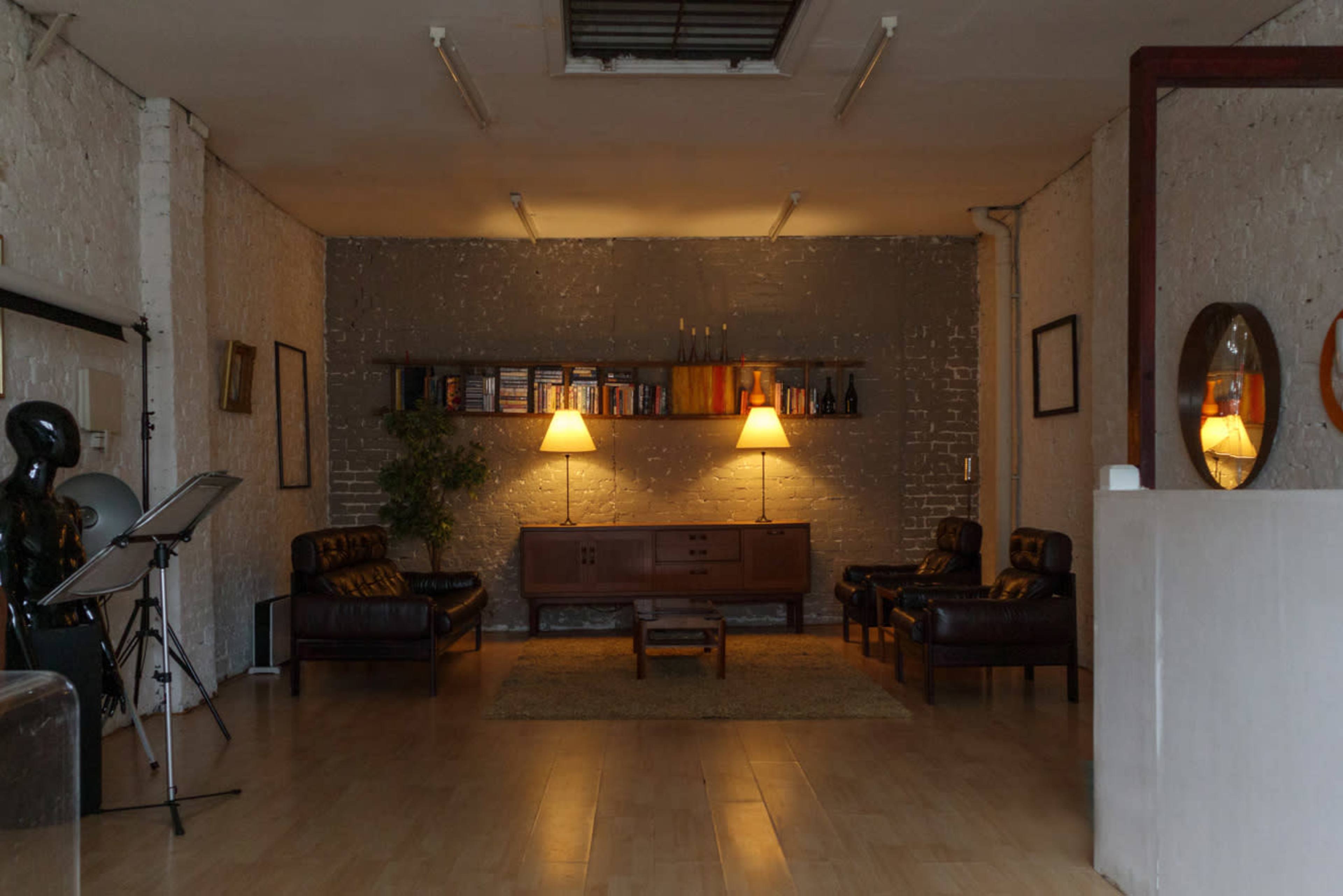 A spacious room features two leather sofas, a low wooden table, and wall-mounted bookshelves illuminated by two lamps.