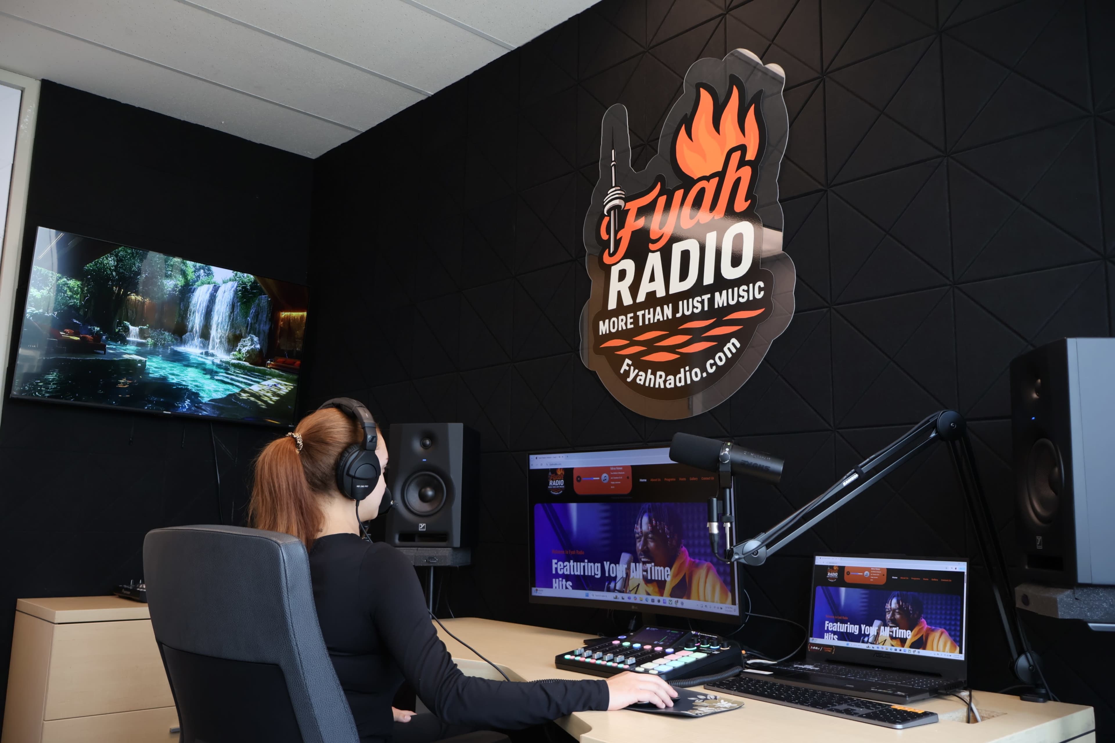 A person in a black outfit operates audio equipment in a modern radio studio featuring a large wall logo and a computer setup.