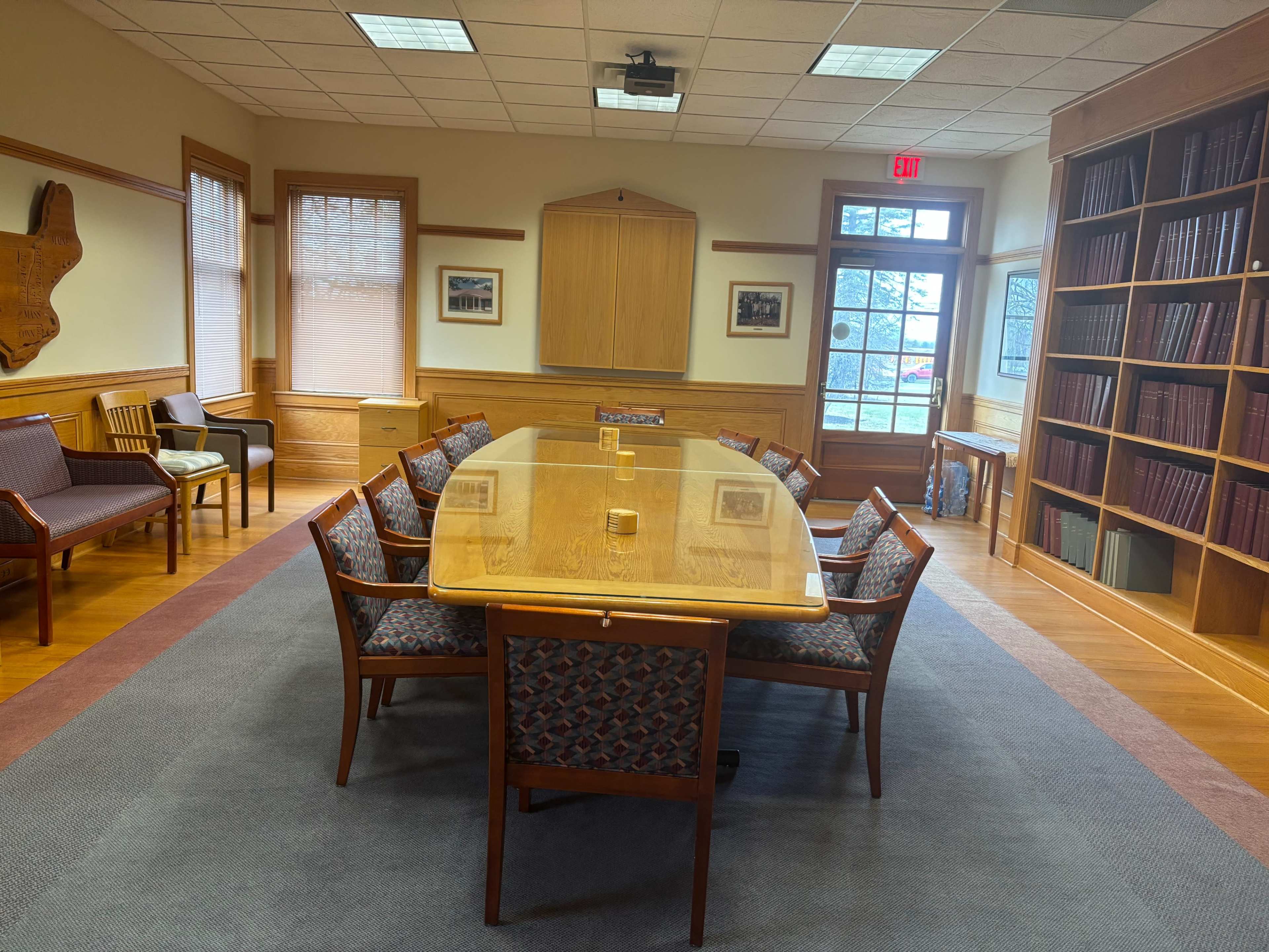 The image shows a small conference room with a large wooden table surrounded by chairs, natural light coming through the windows, and bookshelves along one wall.