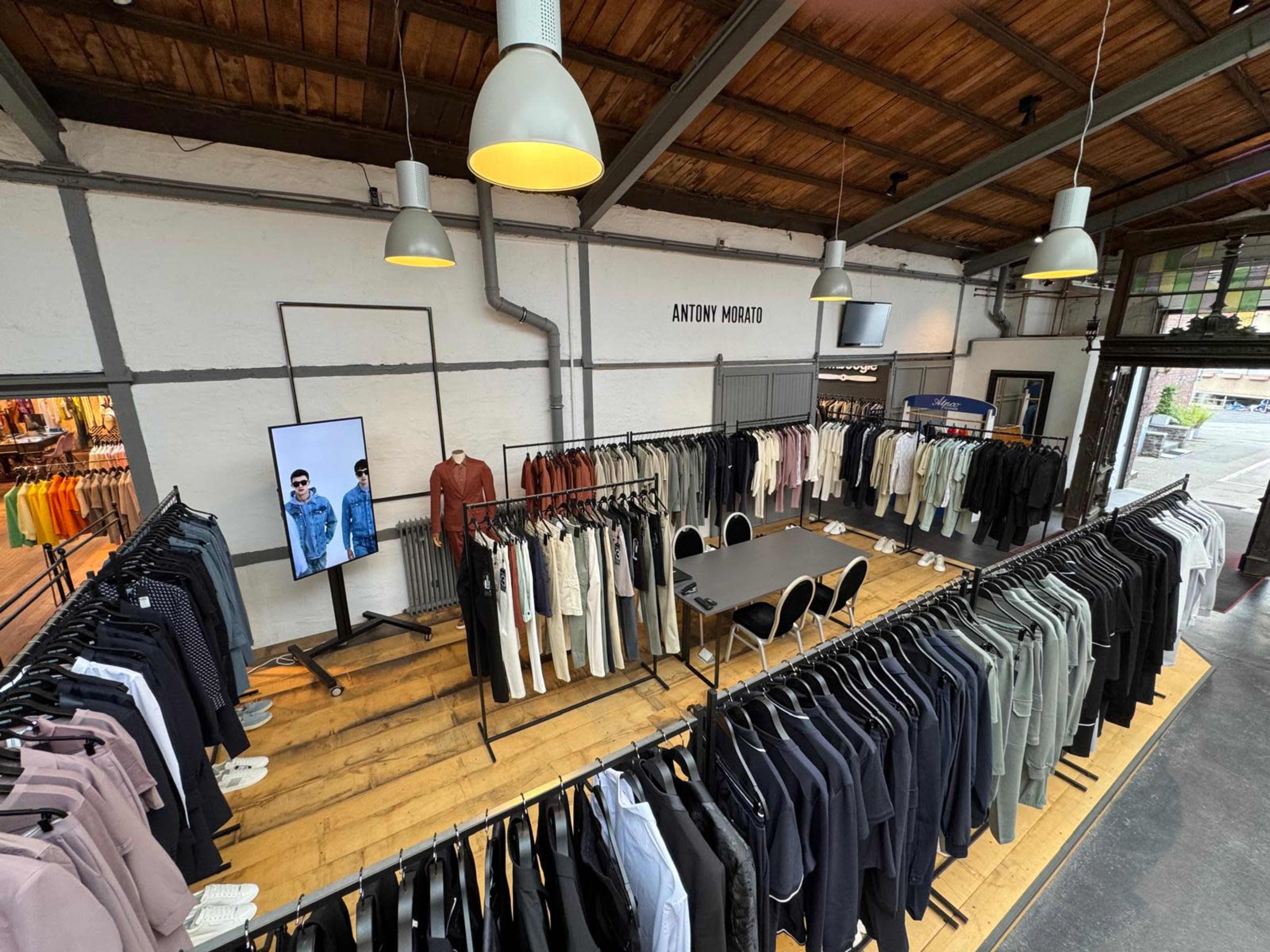 The image shows an interior of a clothing store featuring racks of various men's garments, with a display screen on the side and a seating area in the center.