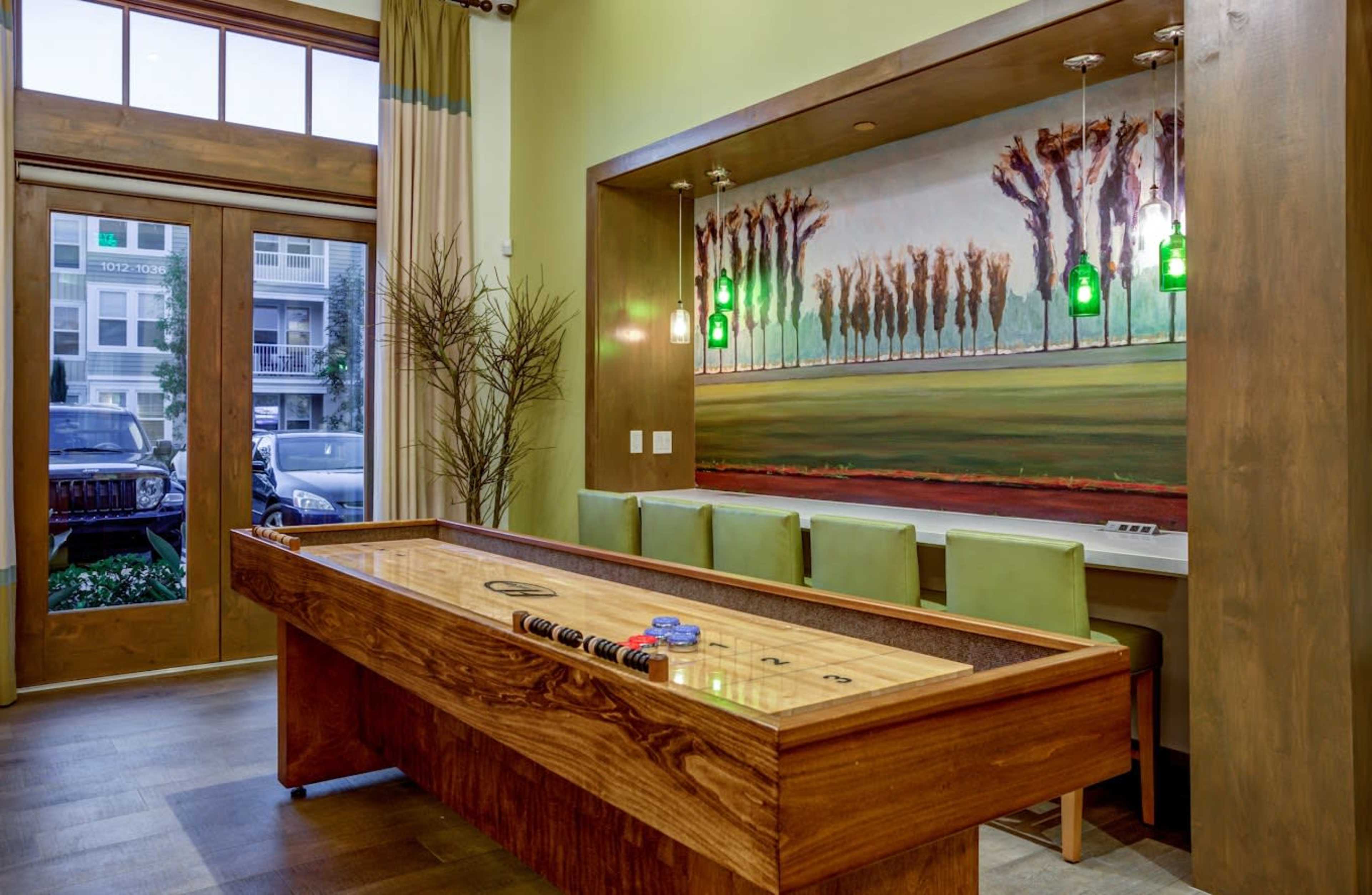A wooden shuffleboard table is placed in a brightly lit room with large windows, featuring a painted landscape mural and a row of green barstools.