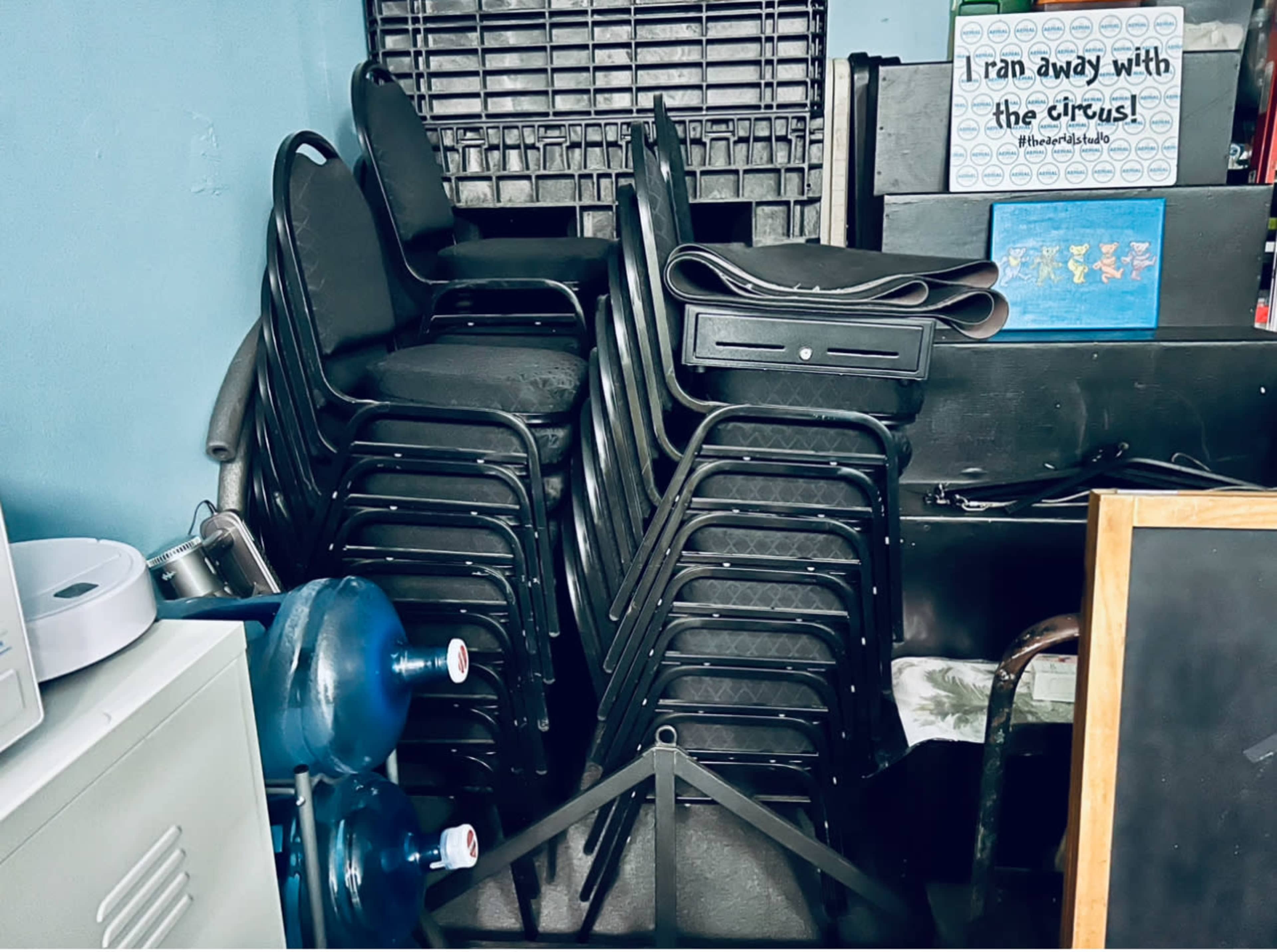 The image shows a stack of black chairs next to a storage area filled with various items, including a large roll and a blue water cooler.