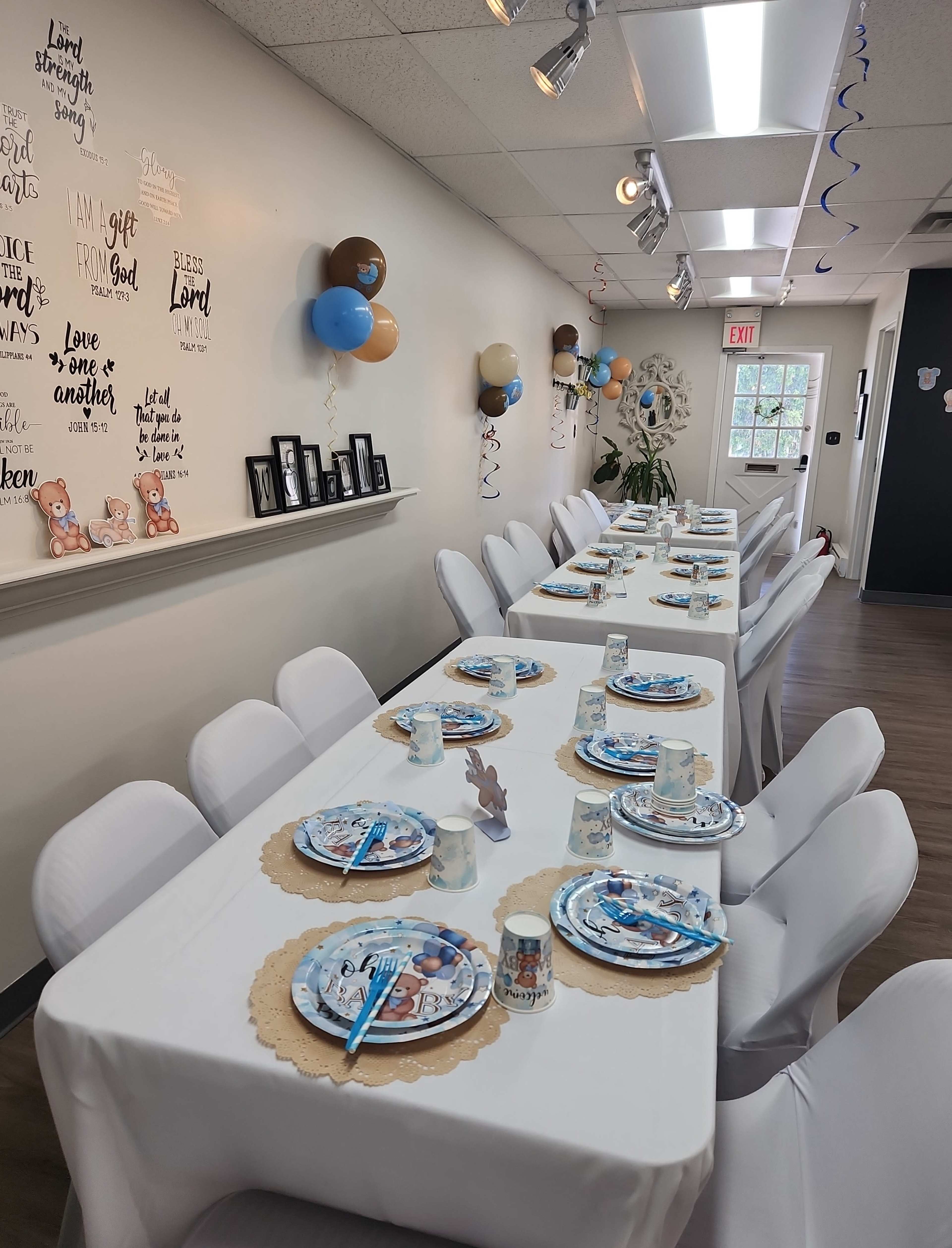 A long table is set with plates, cups, and decorations in a decorated event space featuring balloons and wall art.