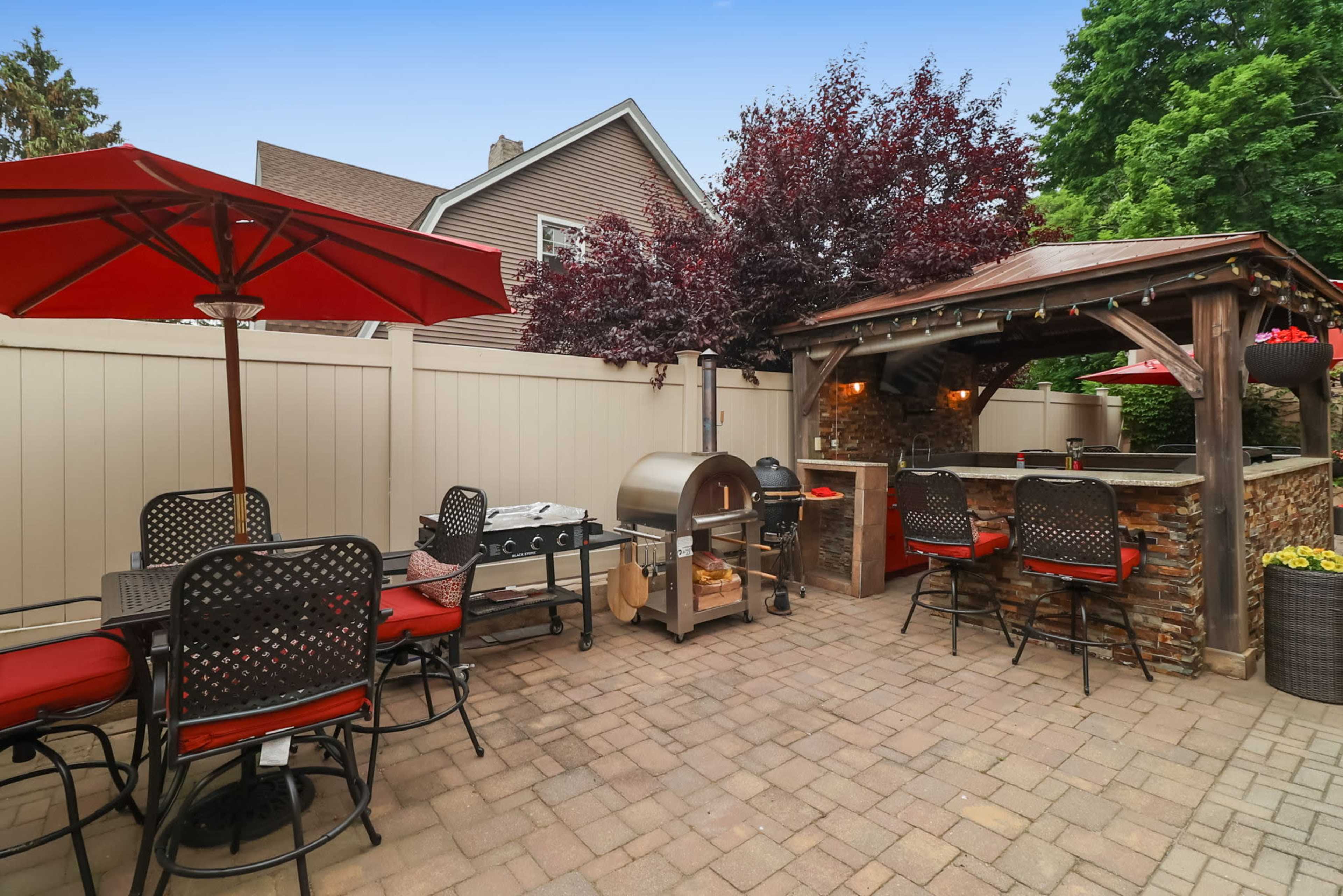 A backyard patio area with a stone outdoor kitchen, a grill, and seating under red umbrellas.