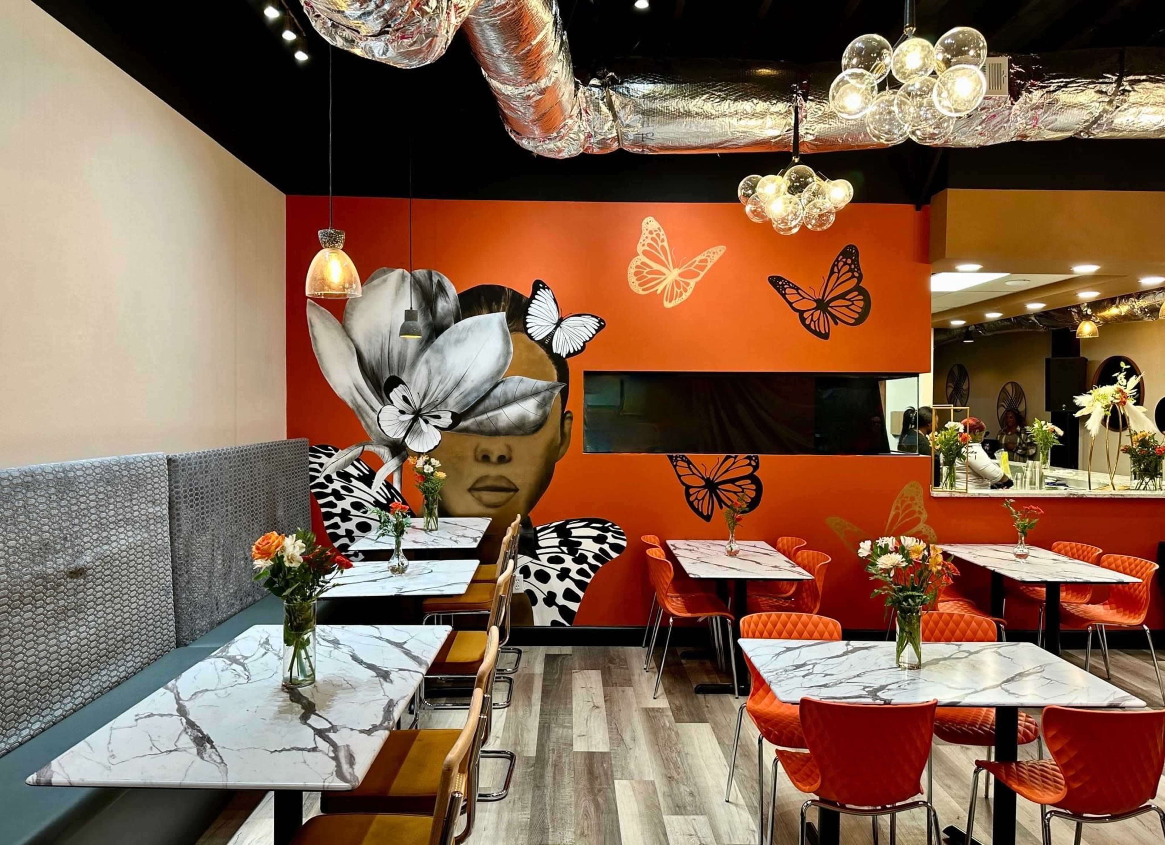 A contemporary restaurant interior featuring marble-topped tables with red chairs, vibrant butterfly decorations on an orange wall, and floral centerpieces on each table.