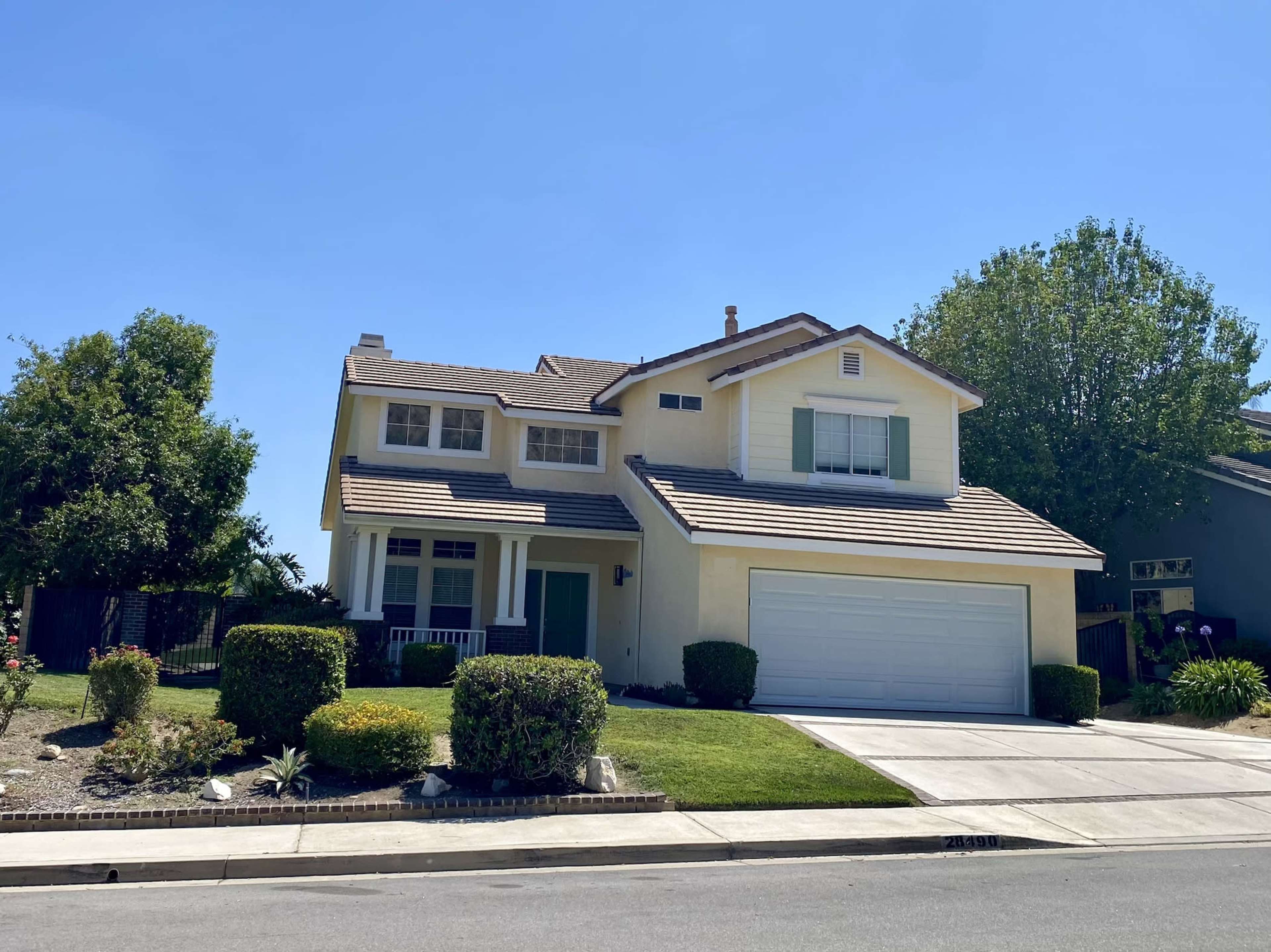 A two-story, yellow house with a brown shingle roof and a two-car garage is situated on a well-maintained lawn.