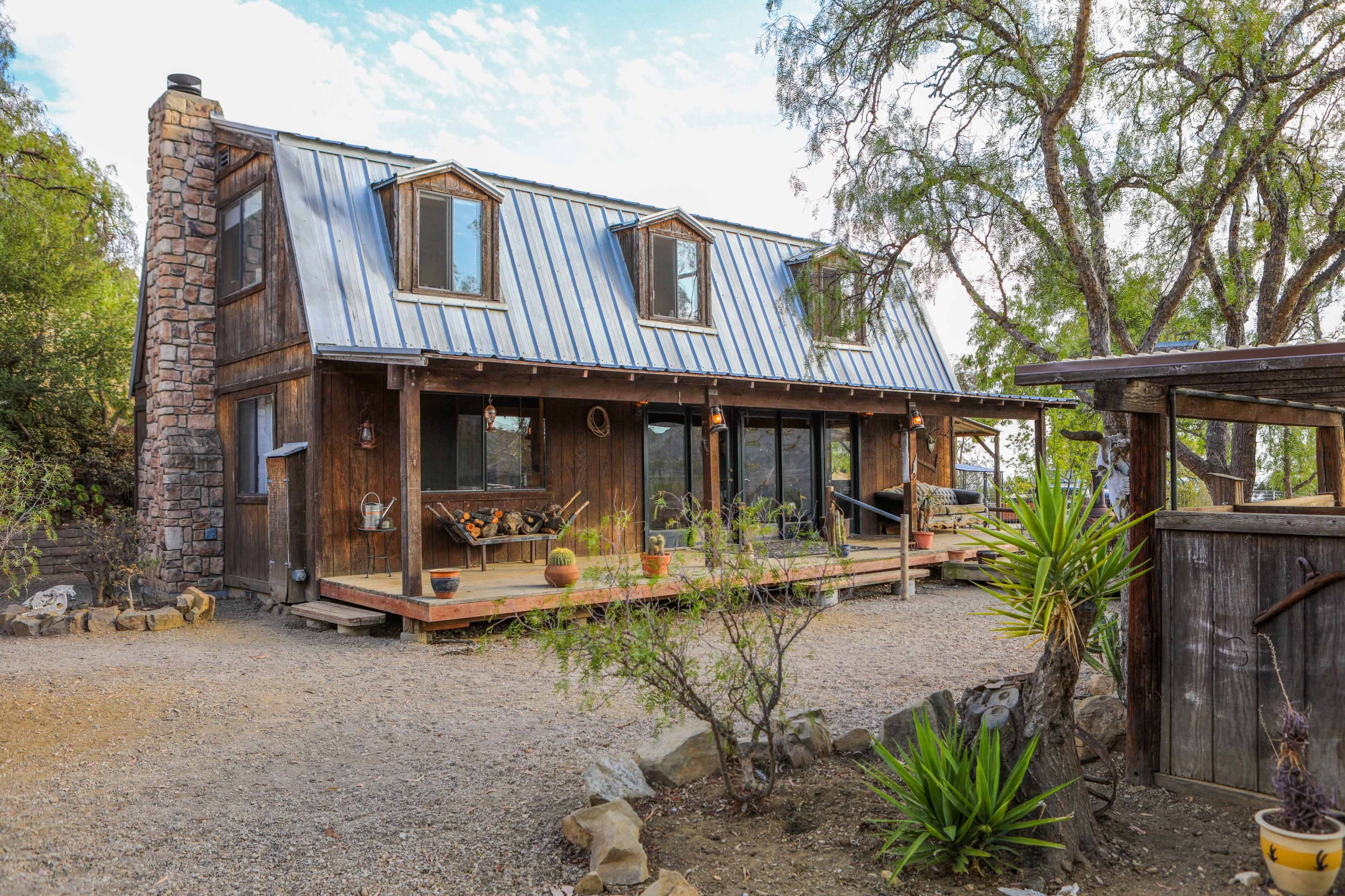 A rustic wooden house with a blue metal roof features multiple gabled windows and a large deck surrounded by gravel and landscaping.