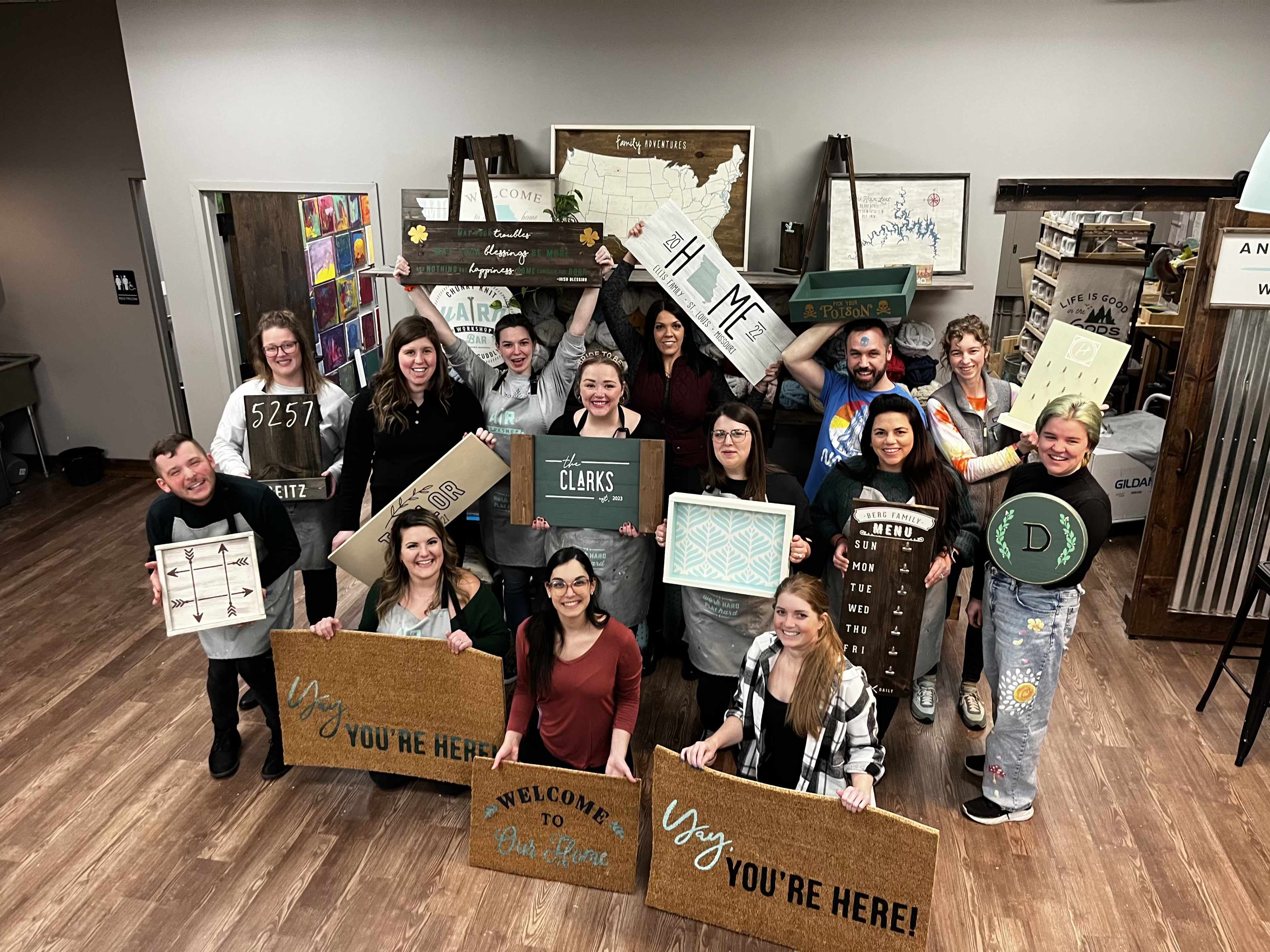 A group of people poses together, displaying various handmade signs and decorations in a workshop setting.