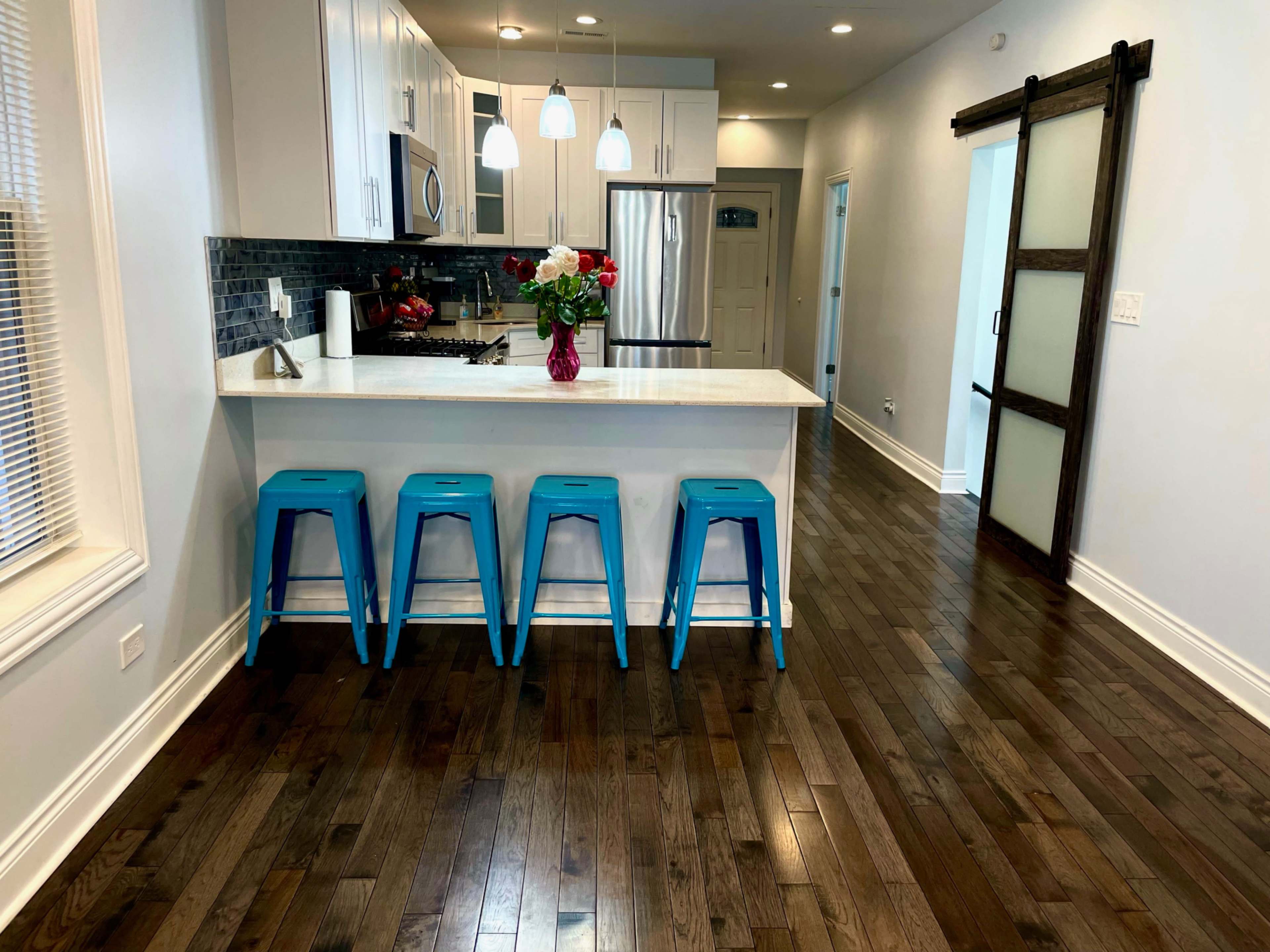A modern kitchen features a white countertop with blue bar stools, stainless steel appliances, and hardwood flooring.