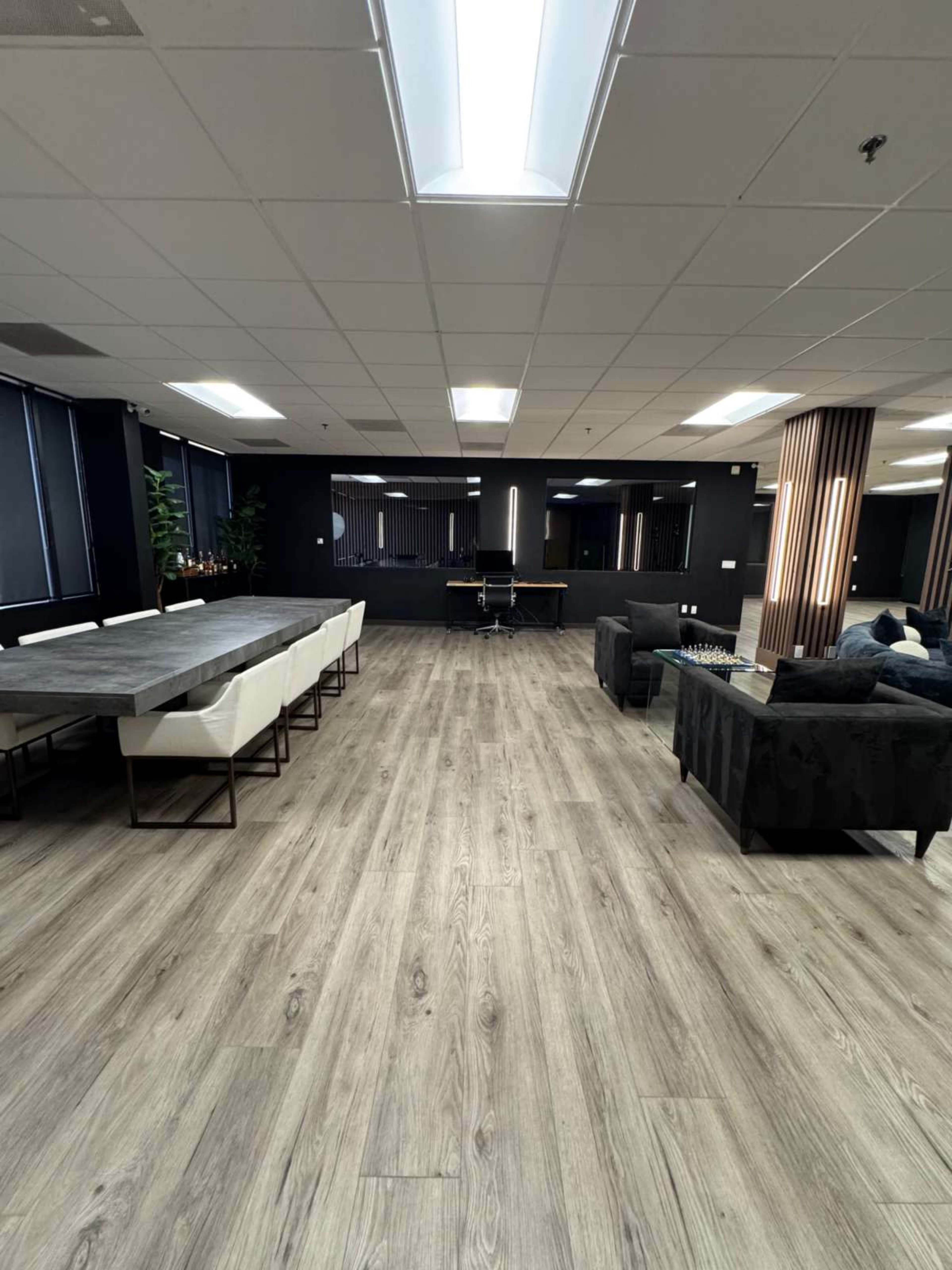 The image shows a spacious, modern office interior with a long dining table, several seating areas, and large windows.