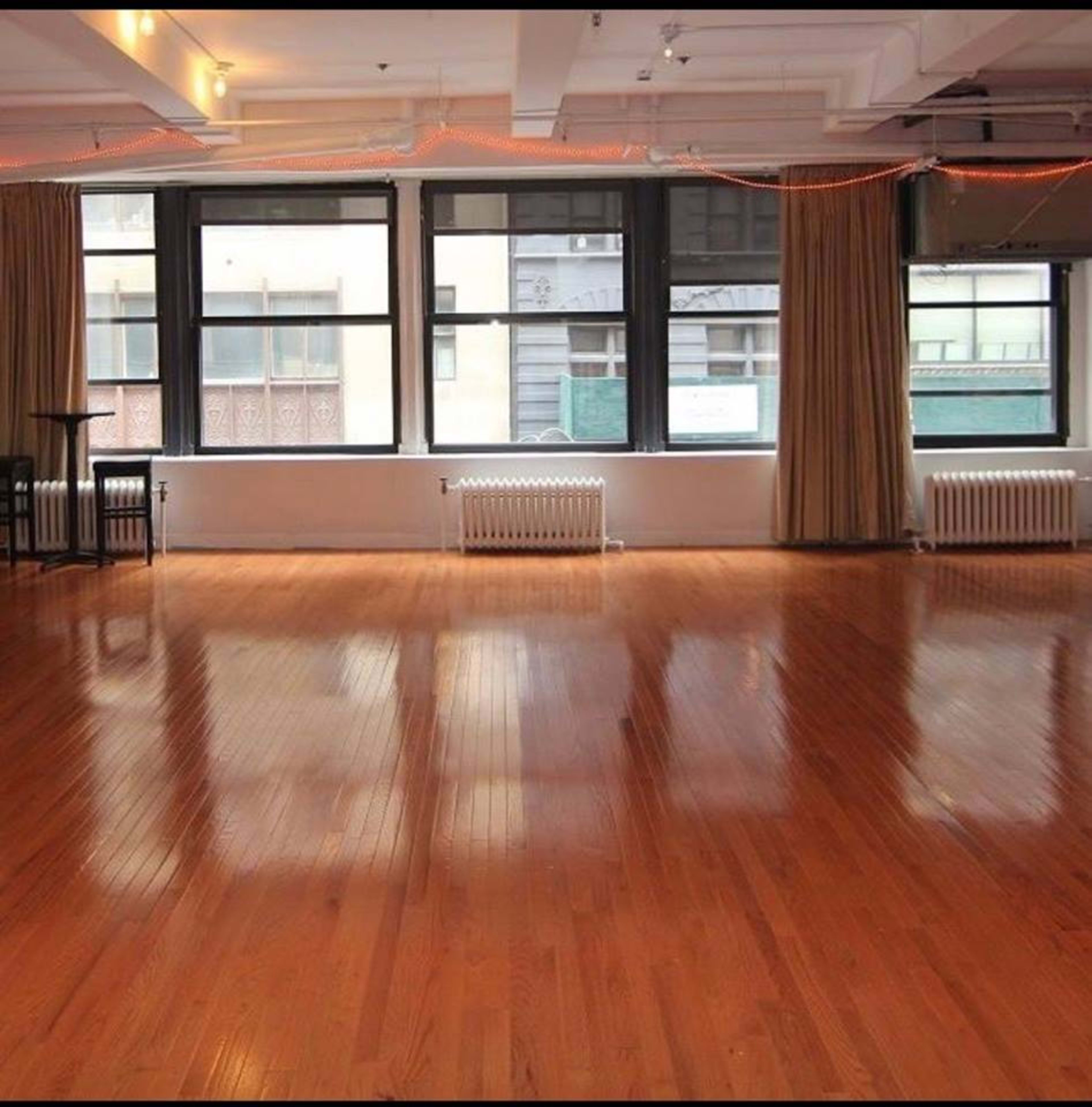 A spacious room with wooden flooring, large windows, and beige curtains.