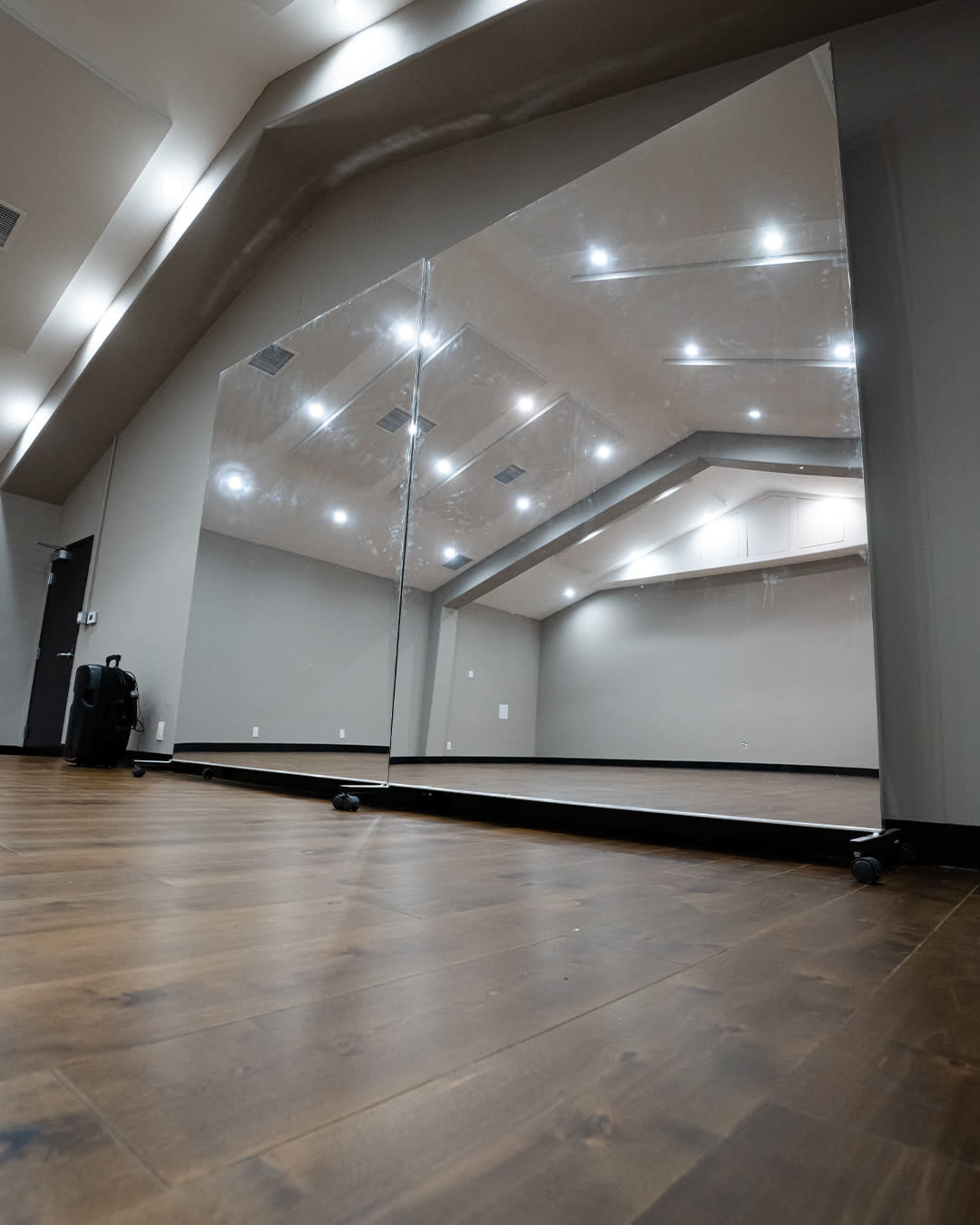 The image shows a spacious room with hardwood floors, featuring a large mirror mounted on wheels and bright ceiling lights reflecting off its surface.