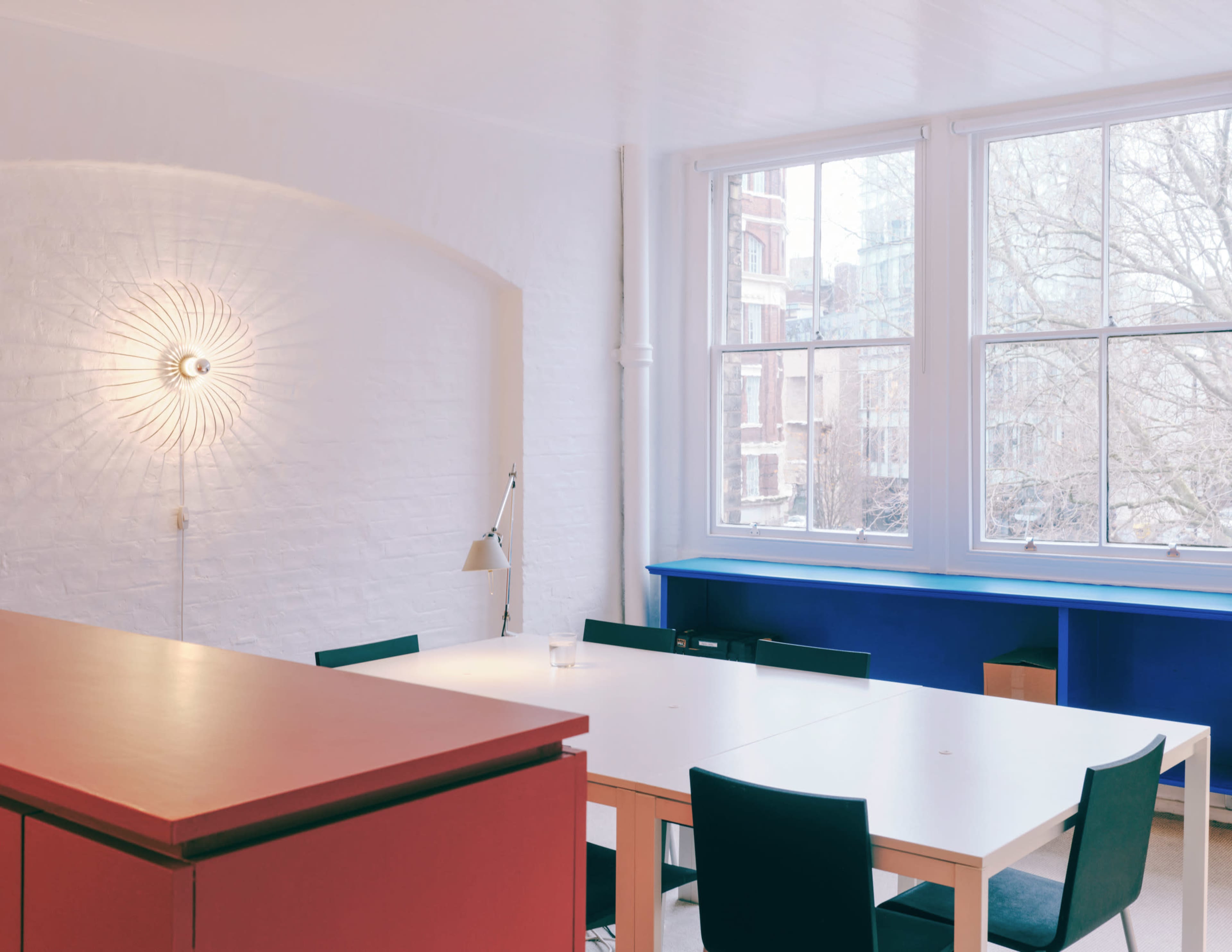 The image shows a modern, bright workspace featuring a large table, black chairs, a red and blue counter, and large windows allowing natural light to enter the room.