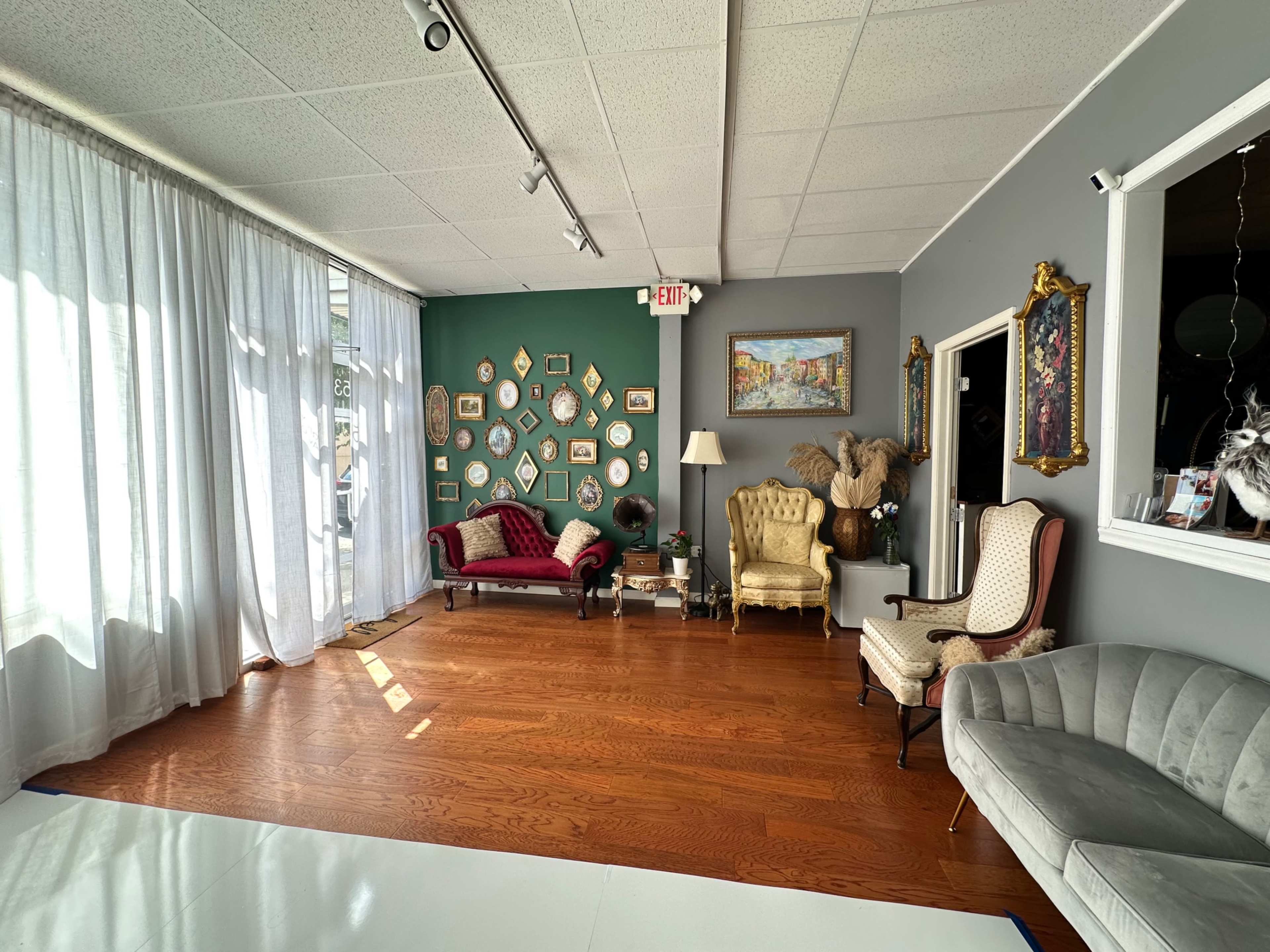 A room with hardwood floors featuring two upholstered chairs, a sofa, a gallery wall of framed artwork, and sheer curtains letting in natural light.