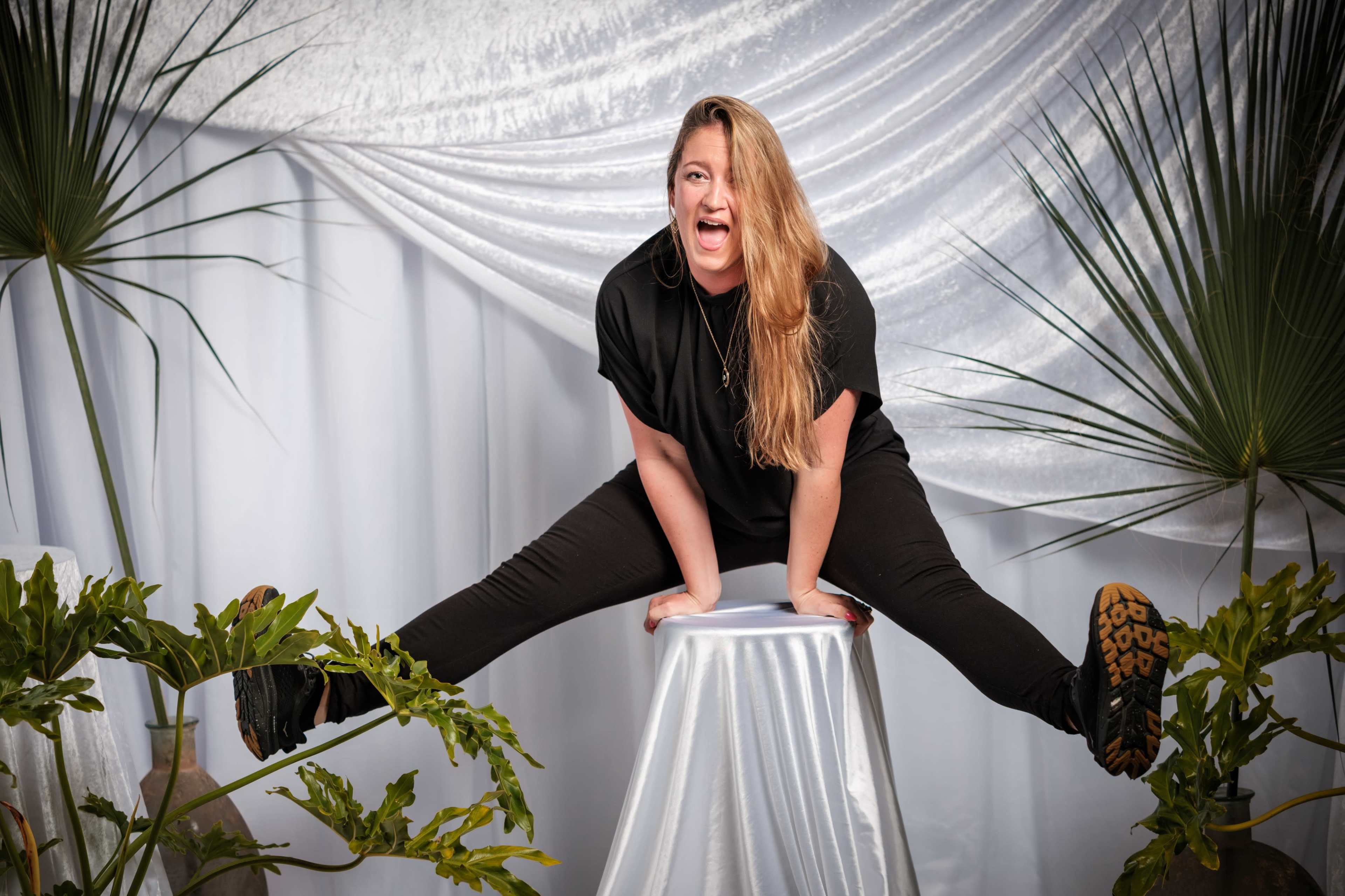 A woman is posing playfully on a white stool with her legs extended in a split, surrounded by green plants and draped fabric.