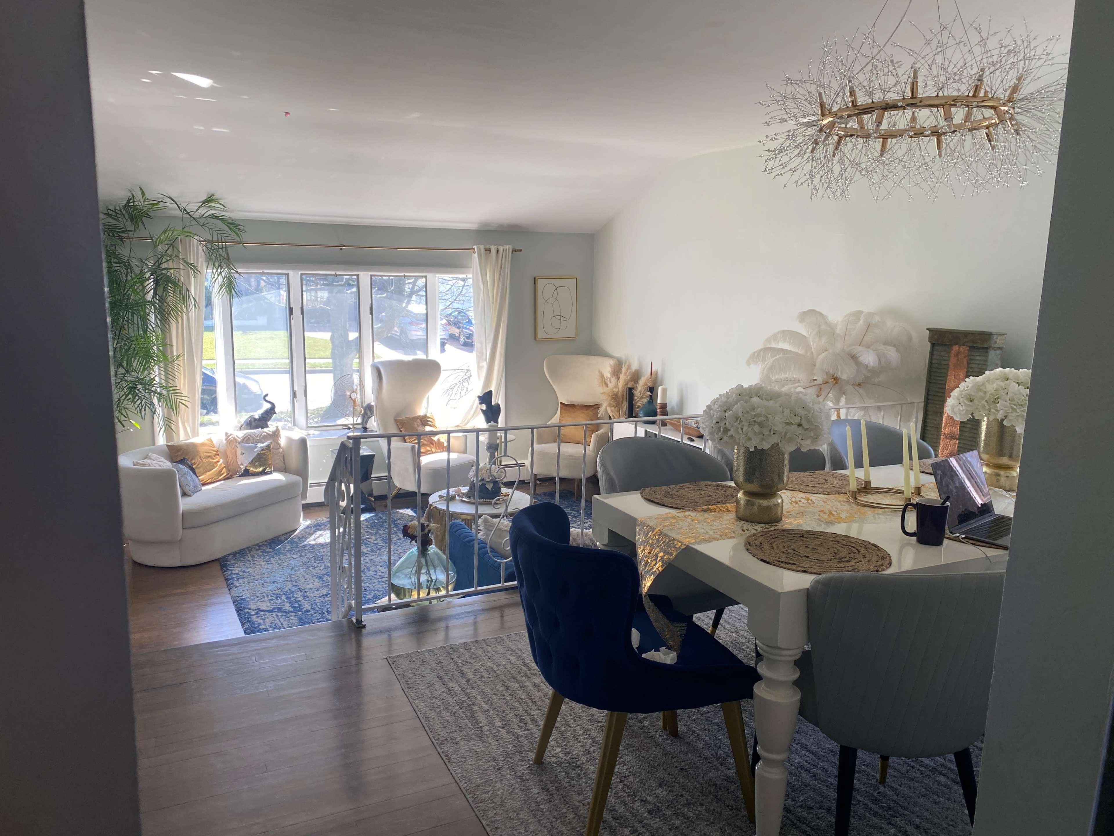 The image shows a bright, open living space featuring a dining table surrounded by chairs, a cozy seating area, and large windows letting in natural light.