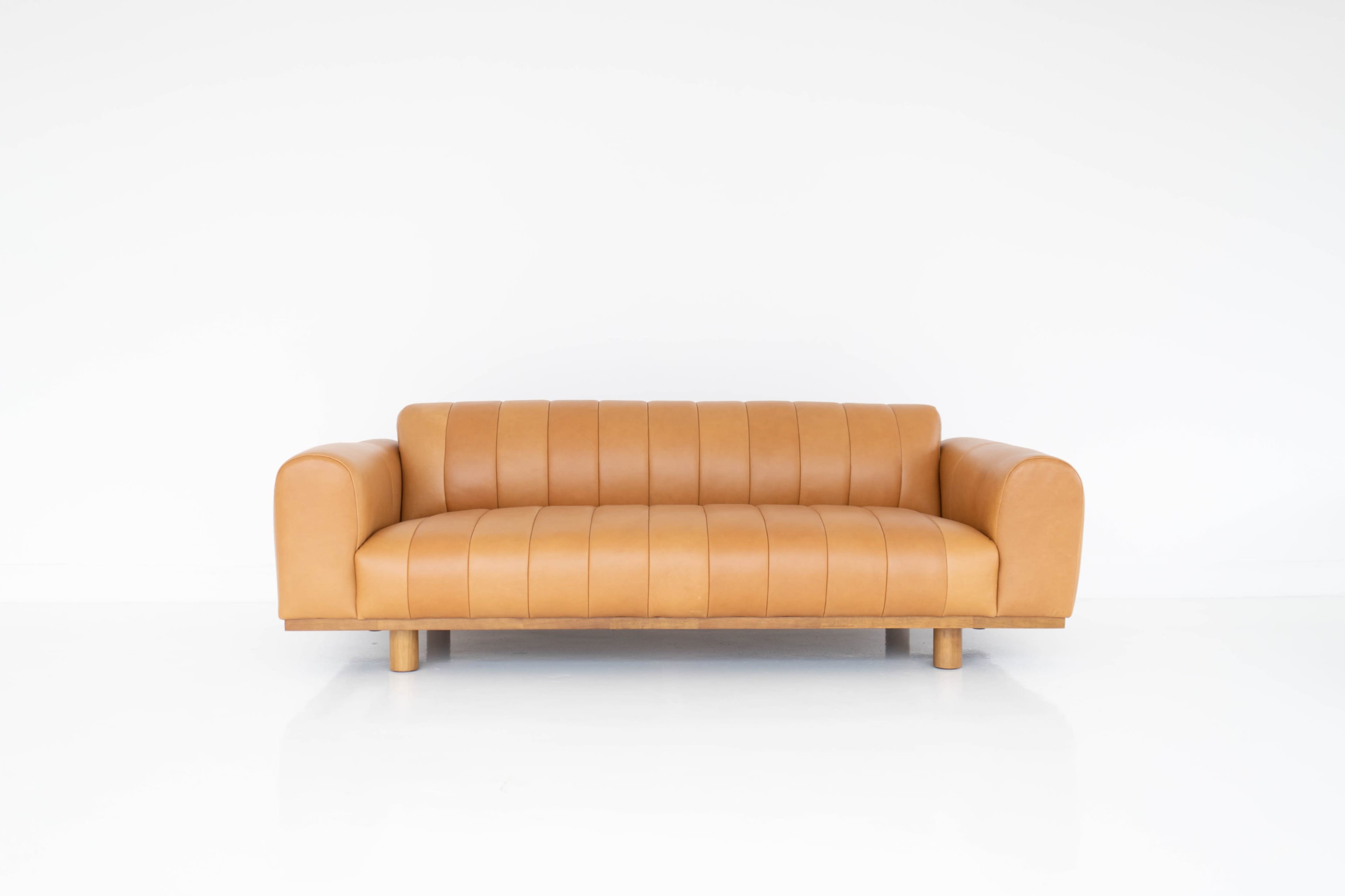 A tan leather sofa with horizontal stitching and wooden legs sits against a white background.