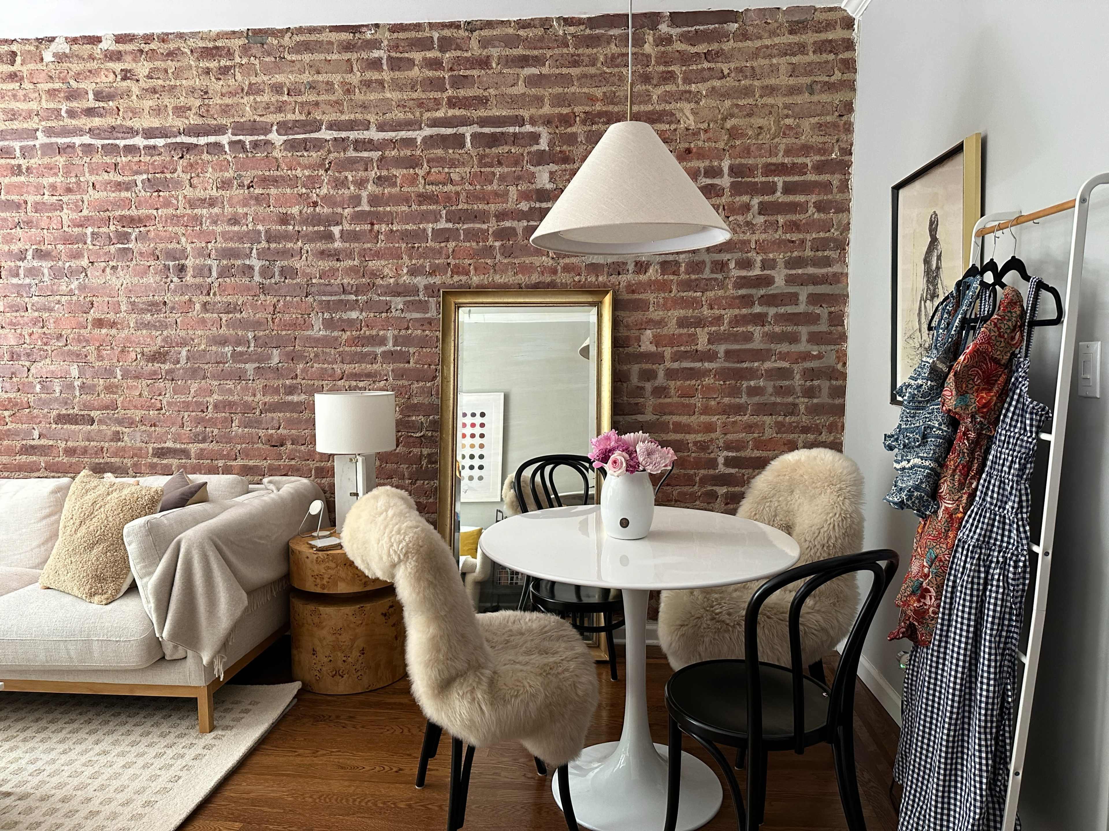 A dining area features a white circular table surrounded by black chairs, set against a brick wall, with a sofa and decorative elements nearby.
