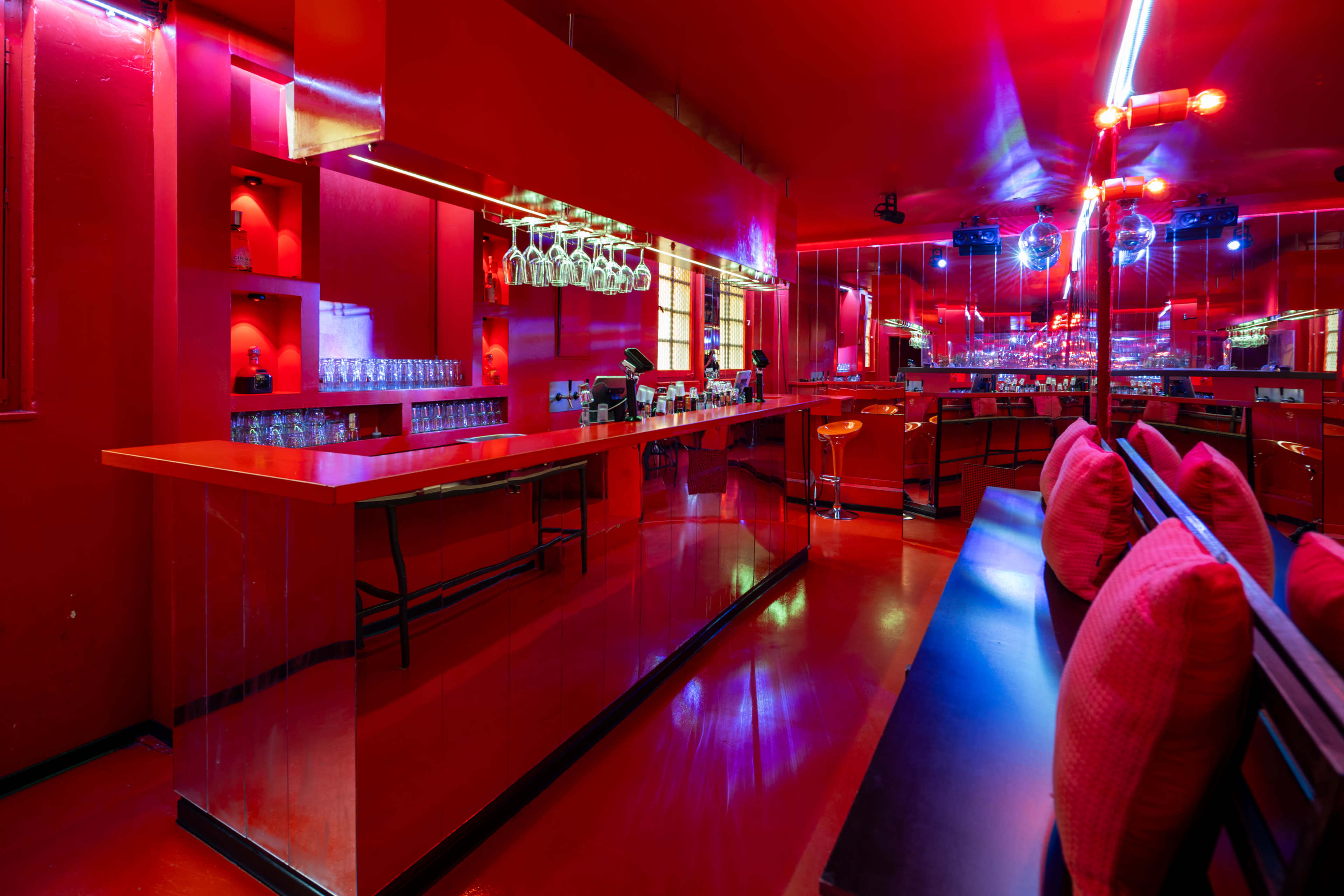 The image shows a brightly lit bar area with red walls, a mirrored bar counter, and seating with cushions.
